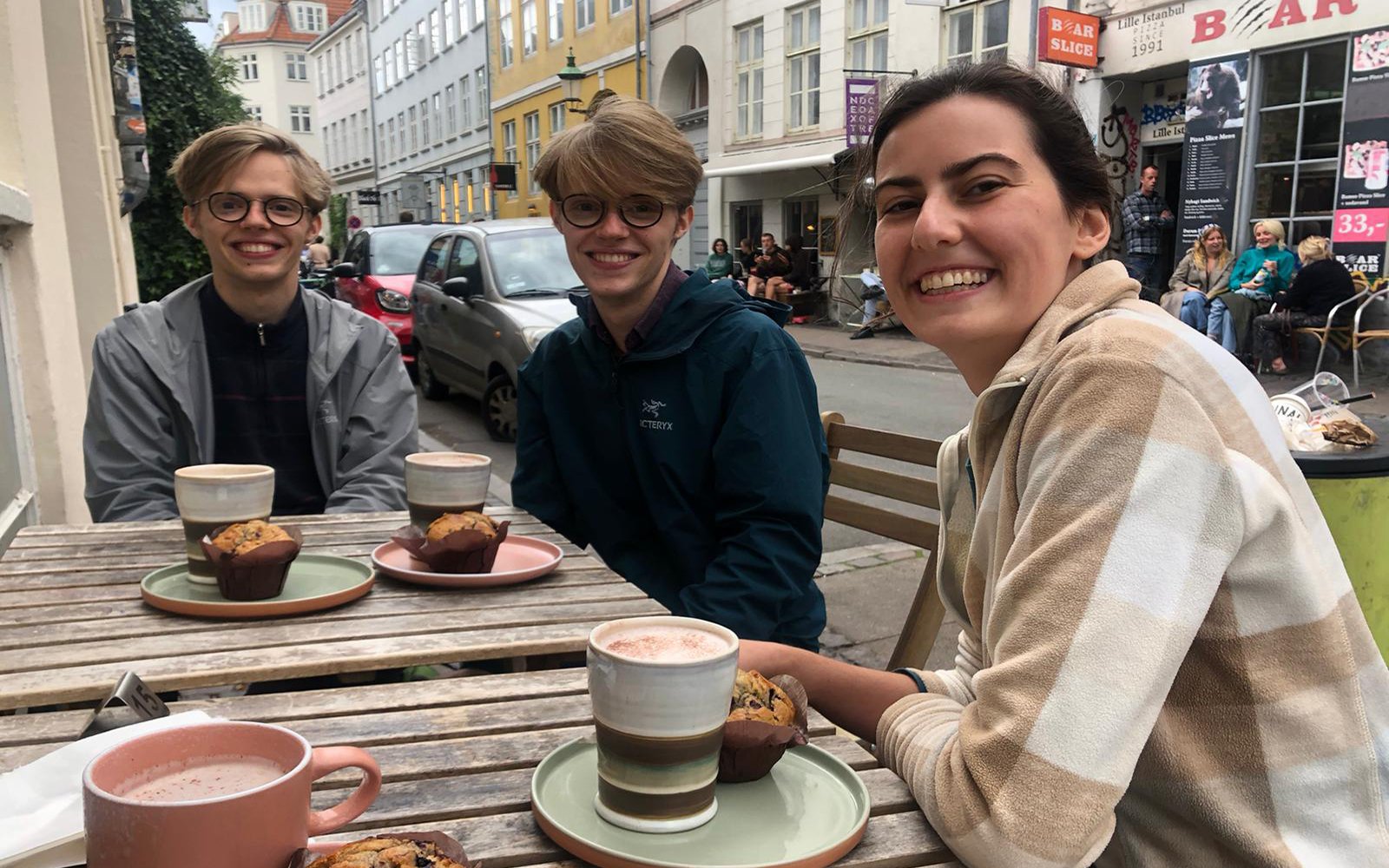 Three students smiling at outdoor table in street downtown, each with a muffin and cup of coffee from a cafe