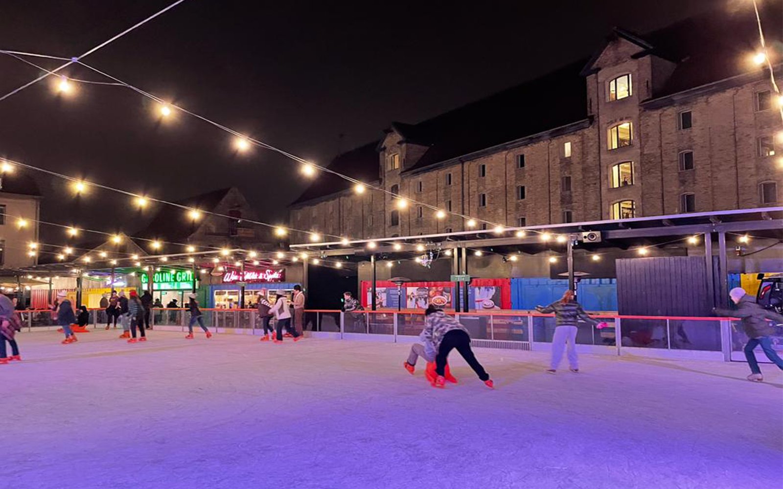 People ice skating on outdoor rink illuminated by string lights next to building at night