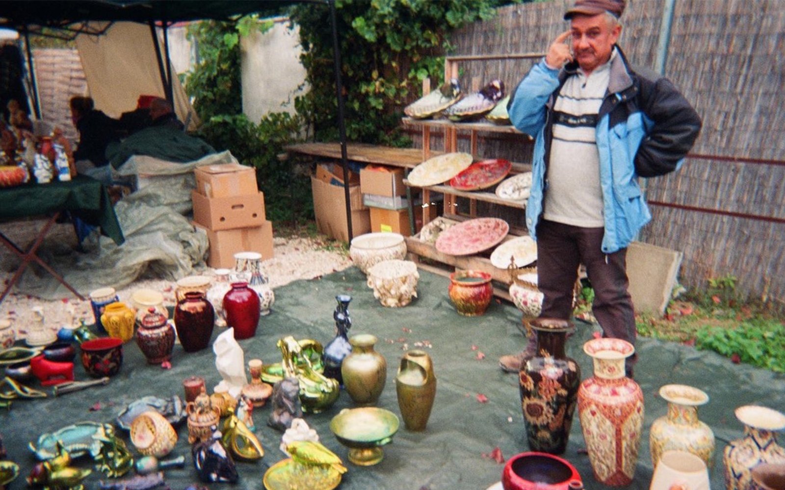 Man standing behind assortment of colorful vases on tarp outside