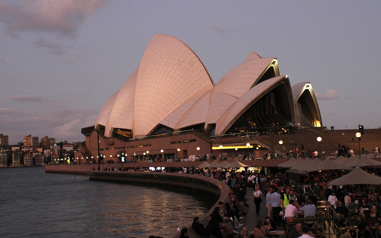 Crowd in front of white building with striking curved segments of roof, overlooking body of water