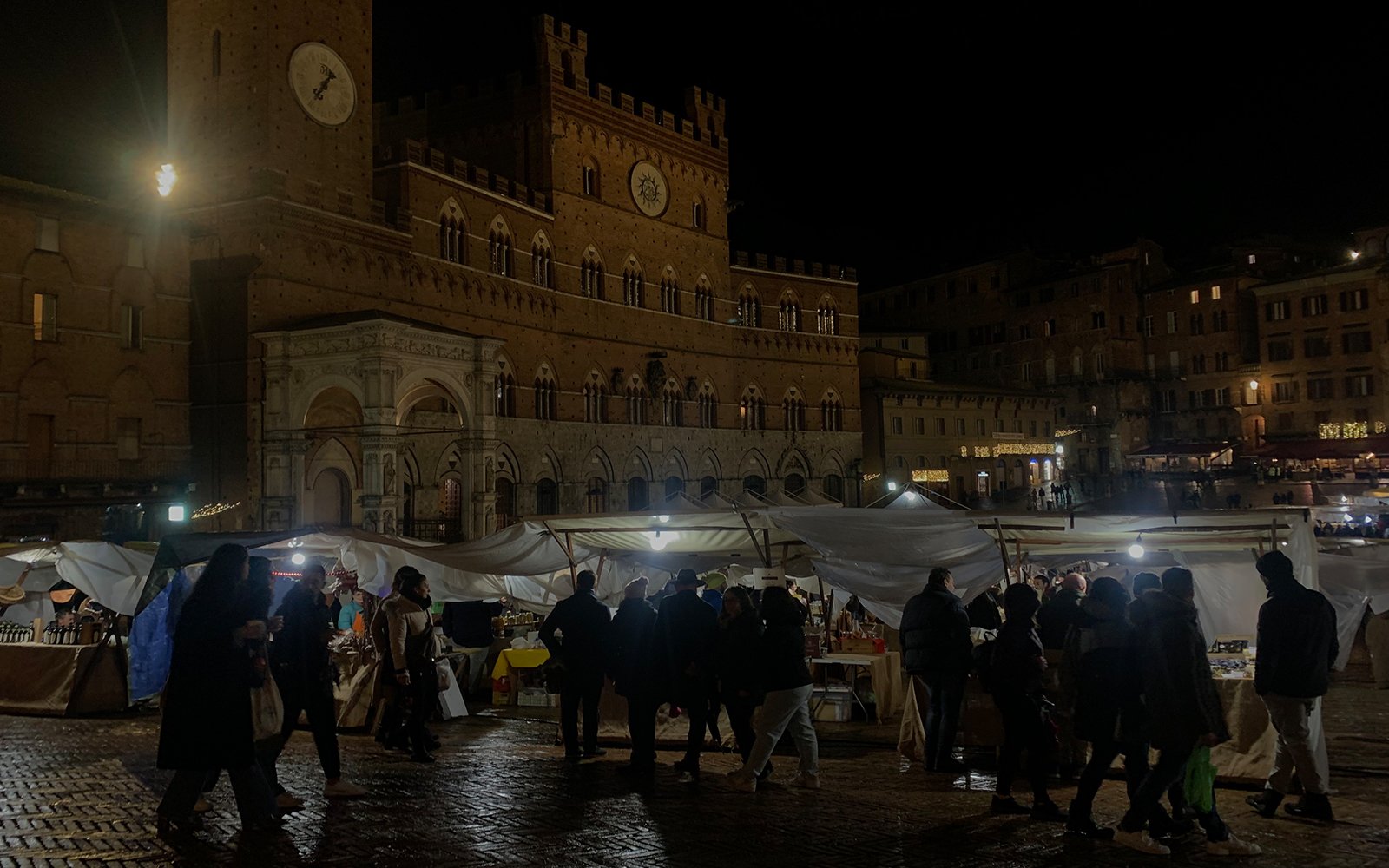 Passersby at nighttime in front of lit outdoor tents for vendors with buildings and clock tower in background 