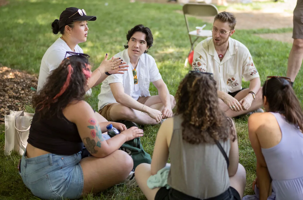 Group of alumni sit on lawn and talk under shade