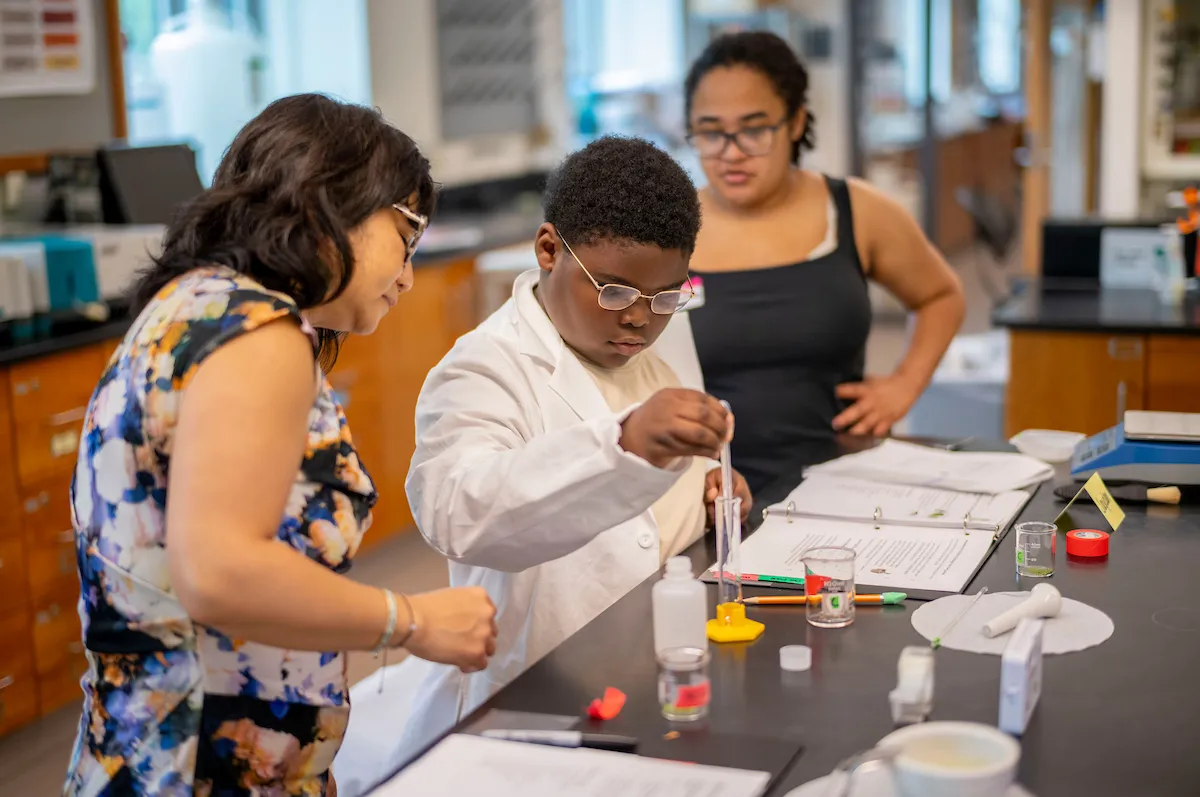 young boy conducts a science experiment alongside a college student and college staff member