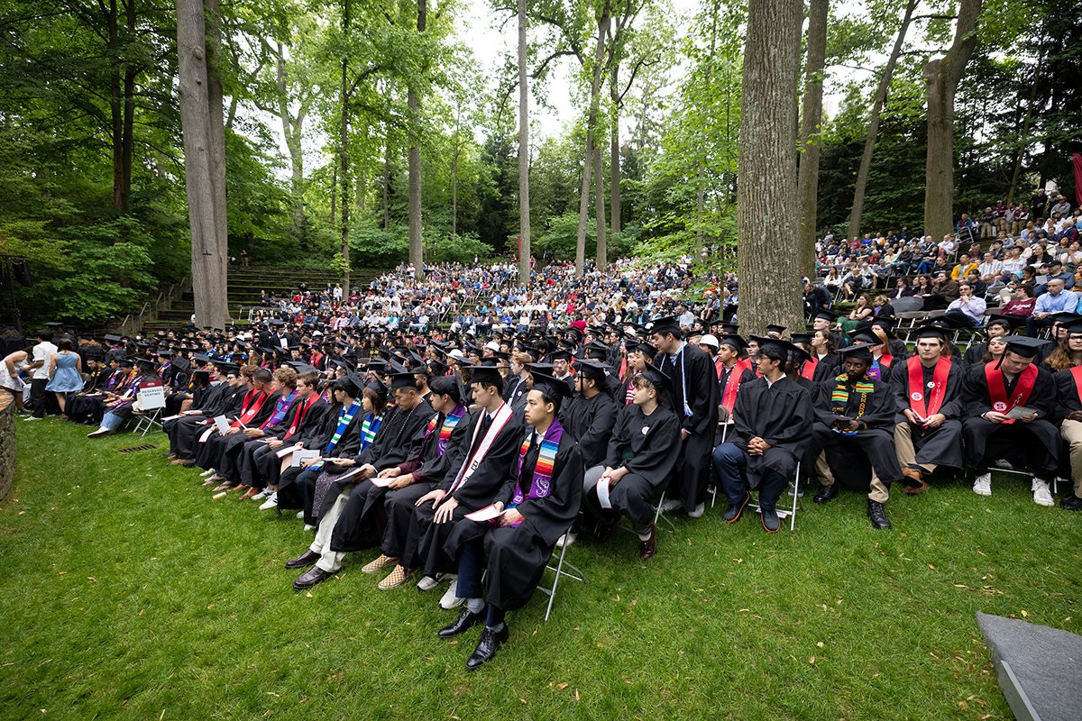 Rows of students wearing caps and gowns sit in outdoor amphitheater