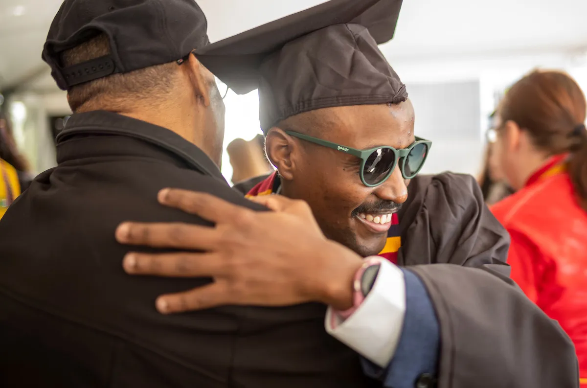 Student in commencement robe and sunglasses hugs man