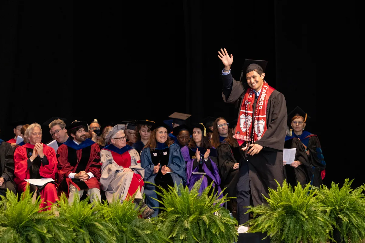 Student waves to crowd while crossing stage in front of faculty