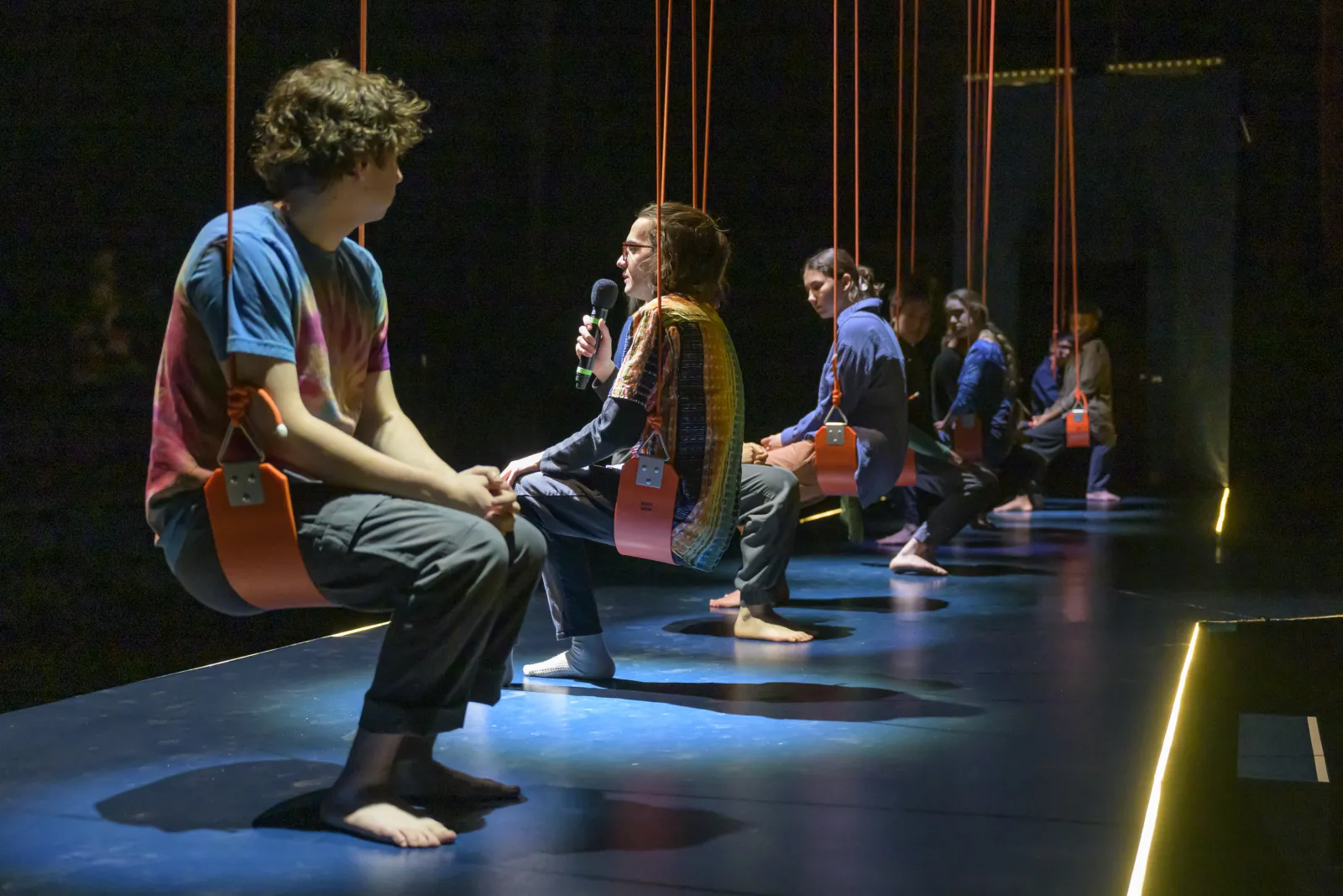 The cast of 32 Very Short Plays About Letting Go. They are seated in swings that have been suspended from the ceiling of the LPAC MainStage. A blue runway is under them, and spotlights from above illuminate each of them. Bryan Arias, a young performer with long hair in a pony tail, speaks into a microphone