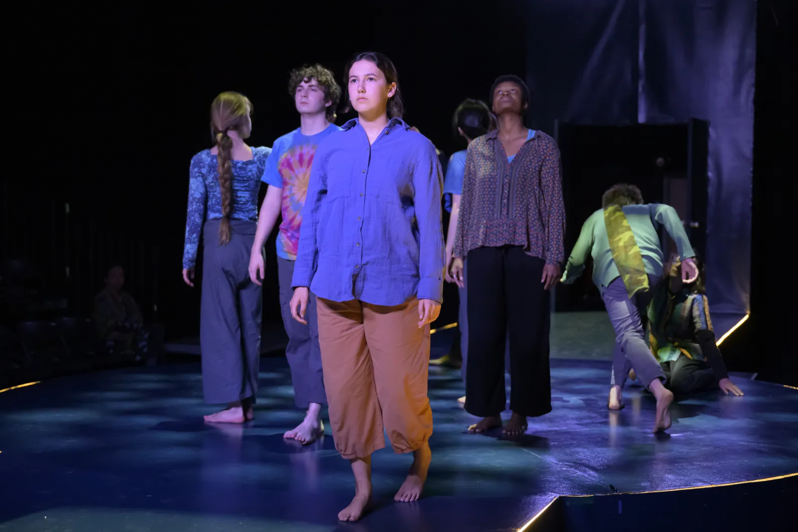 The cast of 32 Very Short Plays About Letting Go. Madison Burch, a performer in a purple shirt, leads a walking dance while the ensemble follows her in the background. The lights are low and moody. 