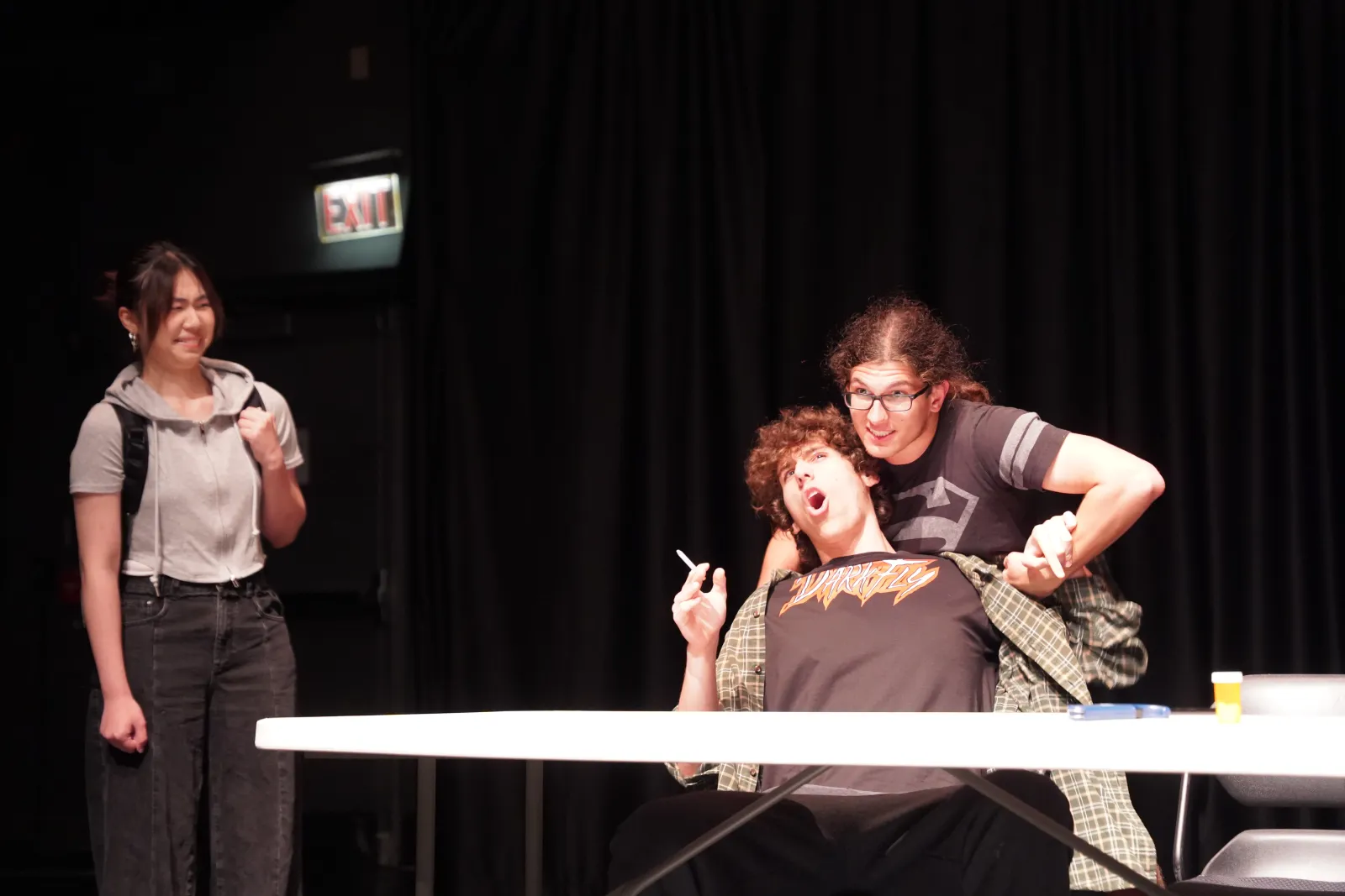 Image from Night of Scenes (Spring 2025). There are three performers, one stands to the far left, and looks on, with their backpack on as if they are about to leave. The other two performers are at a folding table, one is sitting down, but their arms are being pulled back by another performer behind them. It looks like a silly scene
