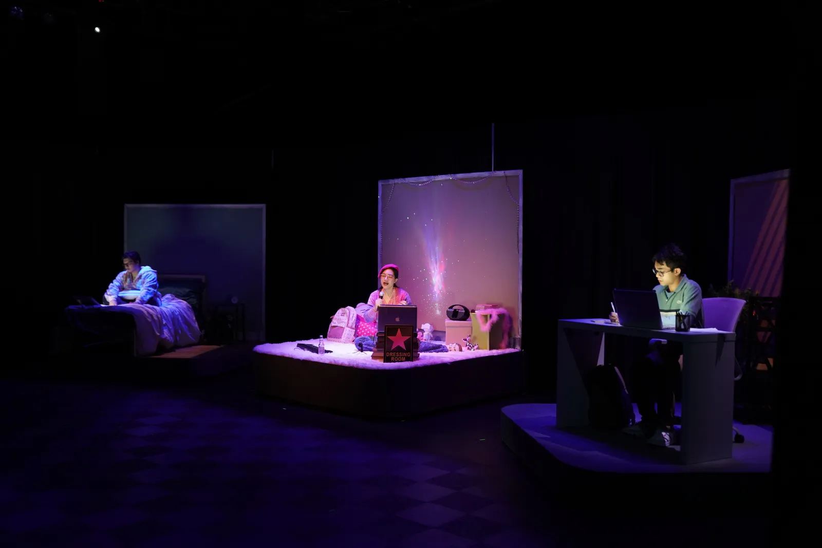 Photo from Speech and Debate. Three students are in their own bedrooms. The far one is dark and illuminated in the blue light of a laptop. A figure can be barely made out. The middle one is awash in pink with a student in a beret. The last room is an office where a student is typing away at their laptop.