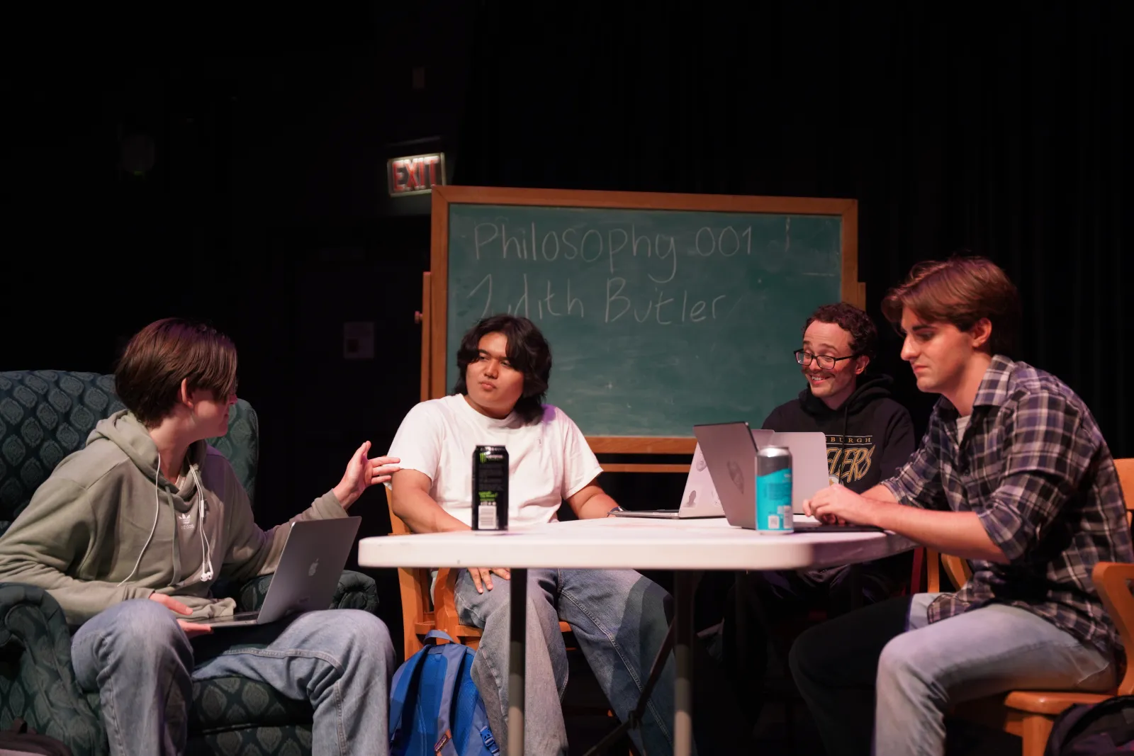 Photo from Night of Scenes (Spring 2025). Four male presenting performers sit around a table in a loose brainstorm situation. Behind them is a chalkboard that reads "Philosophy 001" and "Judith Butler"