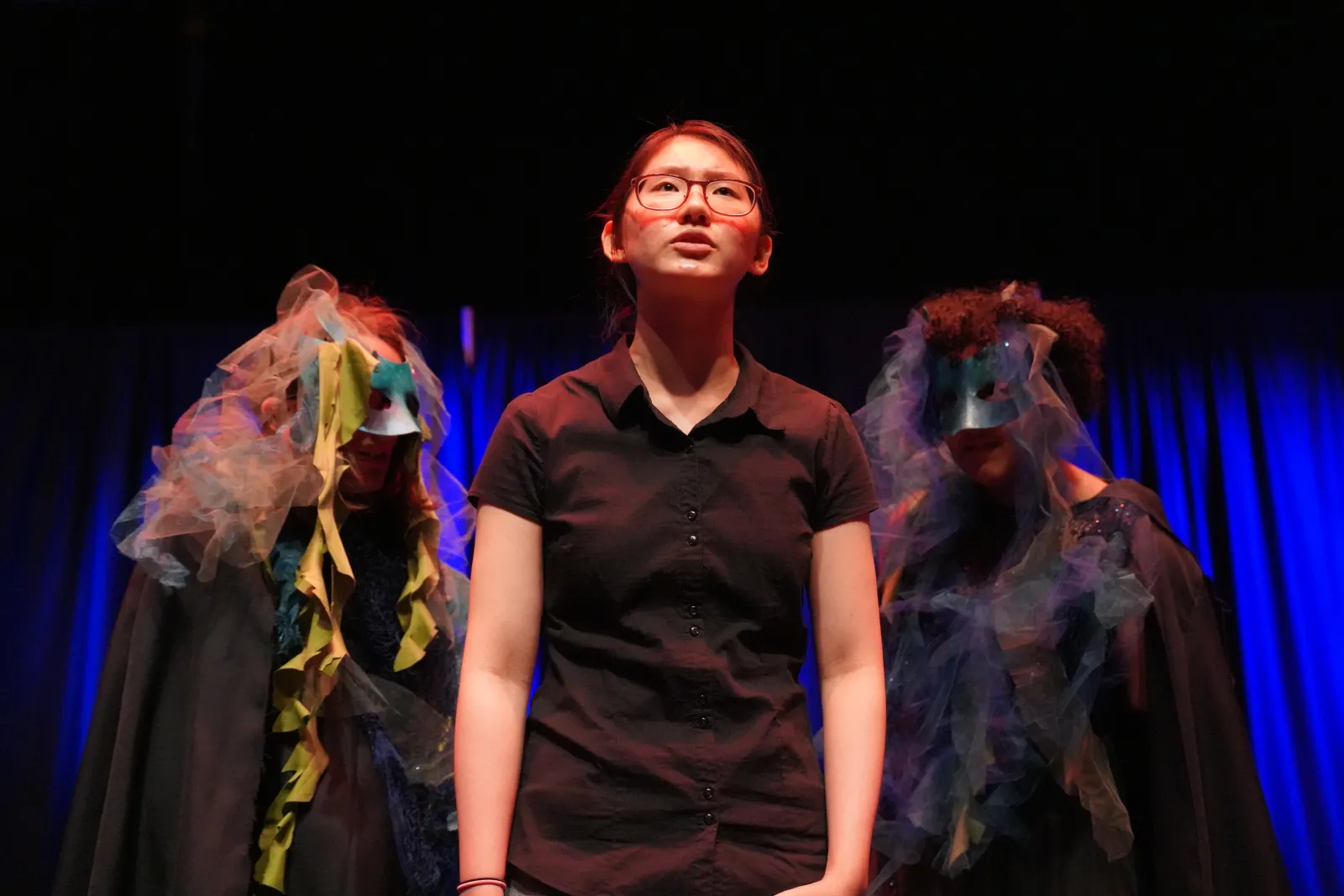 Photo from 2025 Spring Night of Scenes. A performer stands center stage while two masked figured lurk behind them 