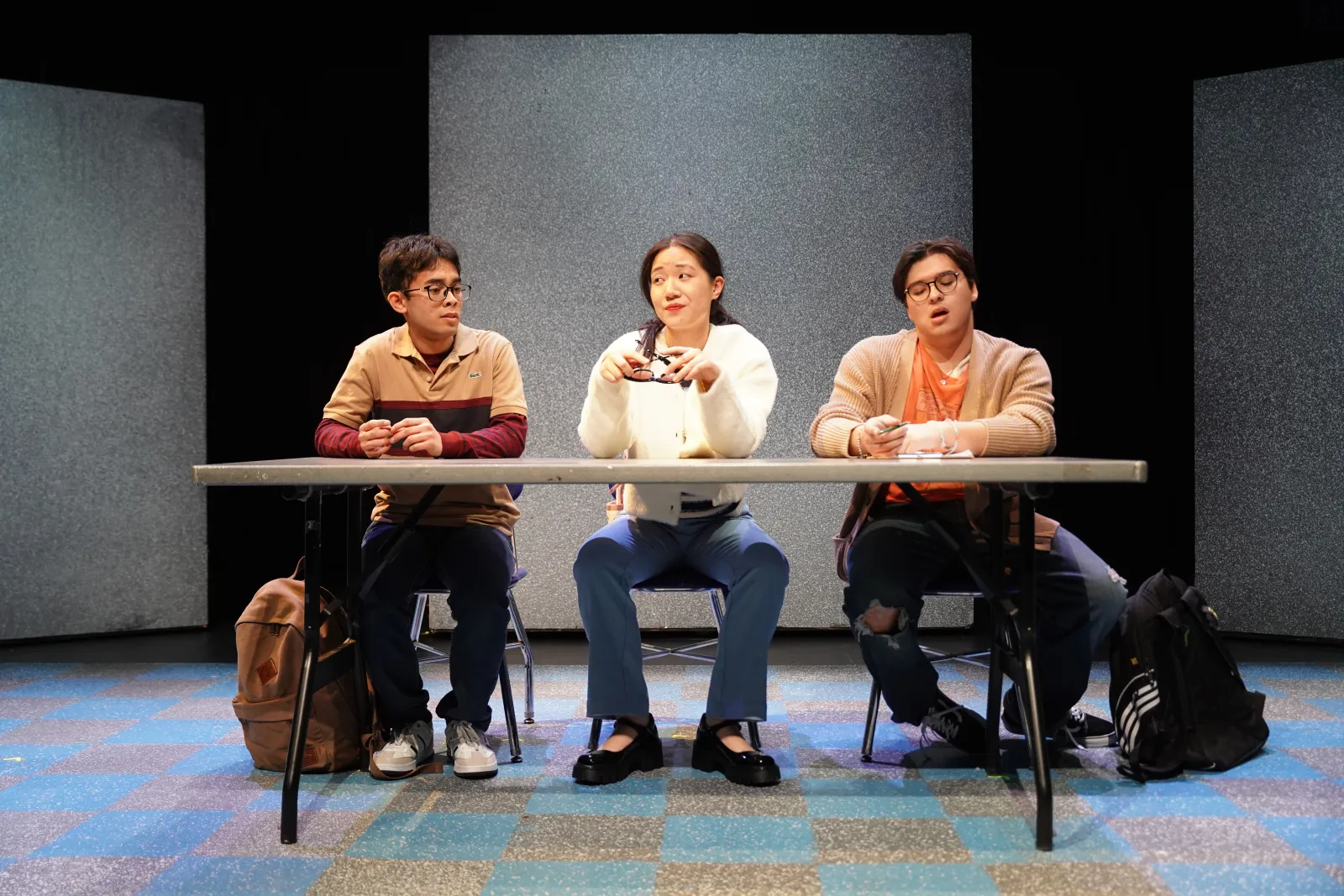 Three performers sit at a folding table. They are dressed like high school teenagers, the student on the left wears a tan and maroon shirt and wears glasses. The student in the middle wears a white cardigan, and is taking off dark sunglasses. The final student on the right looks down and is dressed in a camel toned collared shirt. 