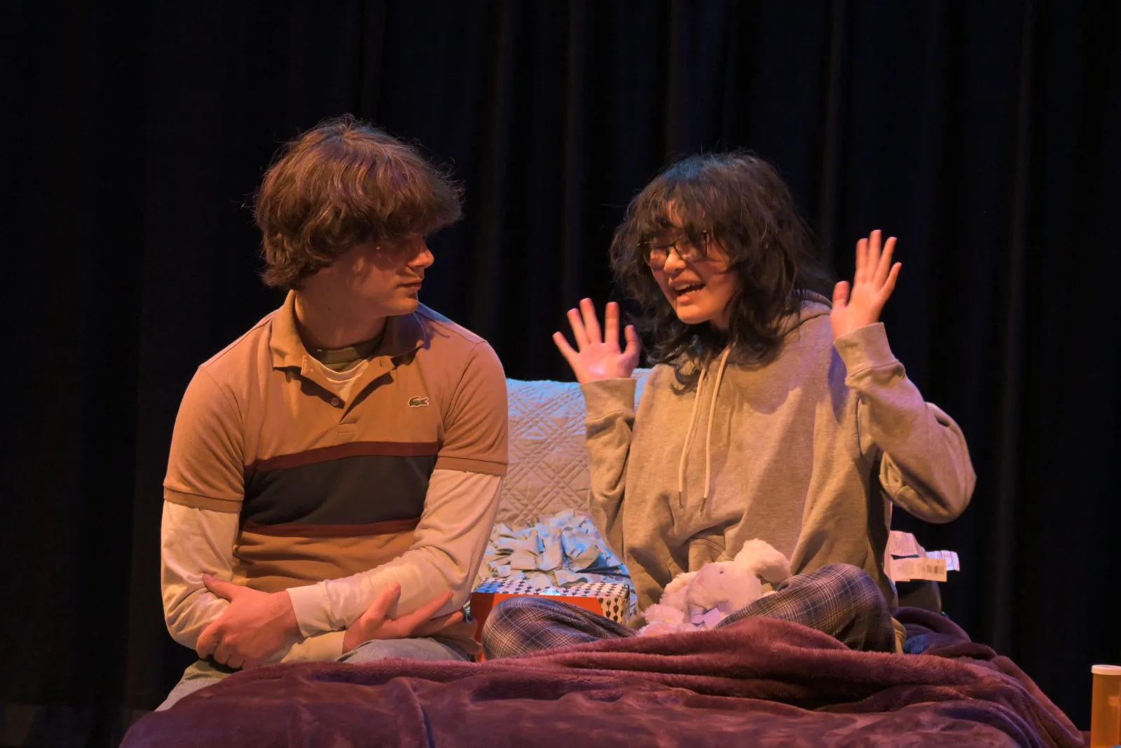 Two performers sit on a bed. Benji Weiss, on the right, has shaggy hair and a polo shirt on. He calmly looks to the individual next to him who has their hands up in the air. They wear glasses and a yellow long sleeve t-shirt. 