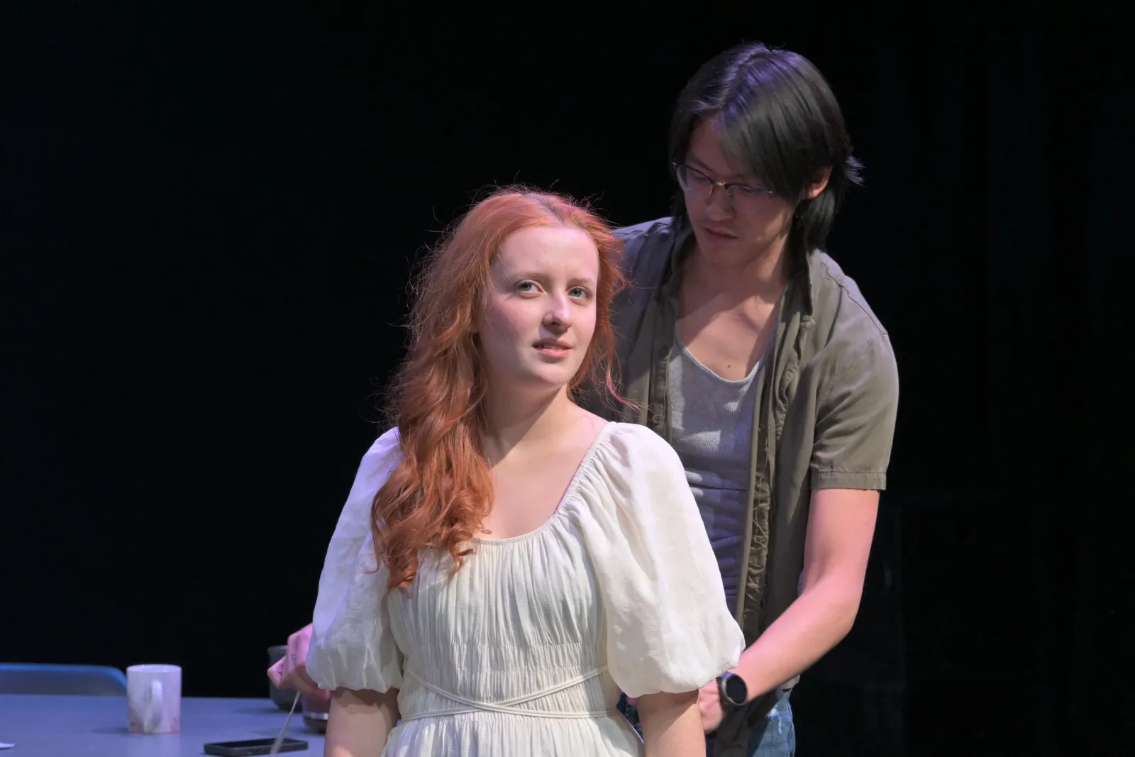 A woman stands downstage wearing a white nightgown. She has red hair and is looking off into the distance. From behind her, a man in a grey undershirt with dark hair looks down and places his hand on her shoulder 