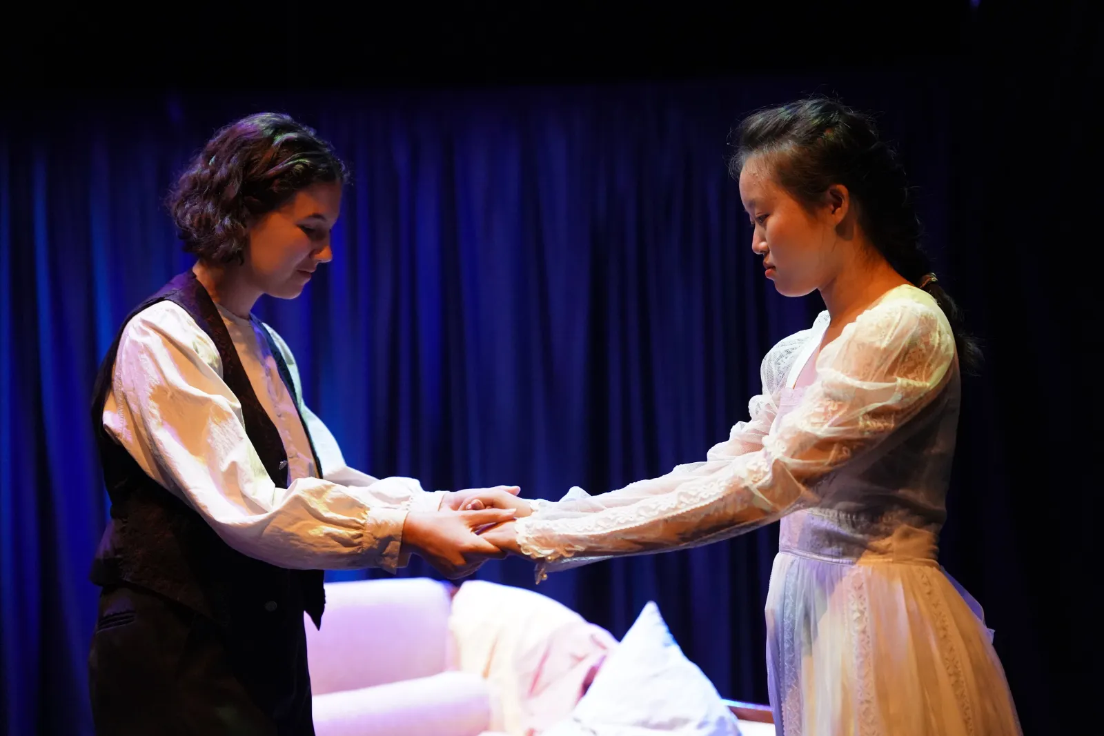 Two performers clasp hands. The performer on the left is wearing a black vest and a white collared shirt, and the performer on the right is wearing a white lace dress