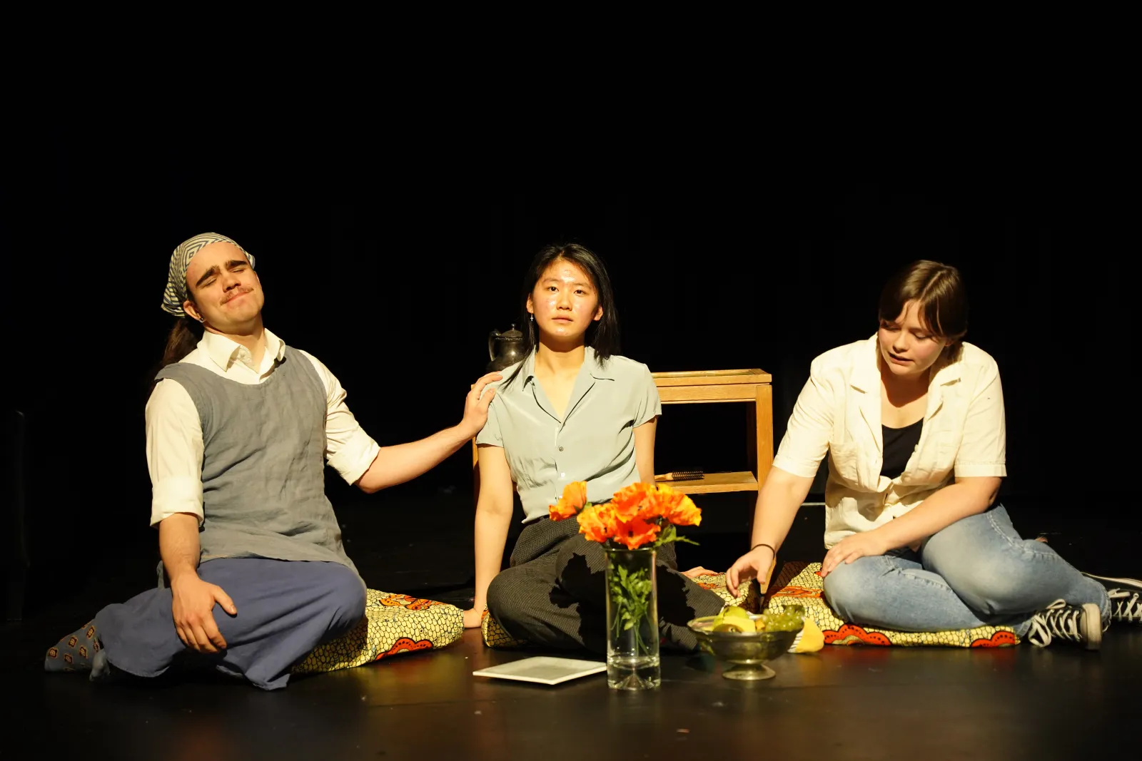 Photo from Fall Night of Scenes. Three individuals sit around a lunch. The performer on the left puts a reassuring hand on the performer in the middle who has a stunned look on their face. The performer on the right is sampling some of the food at the picnic