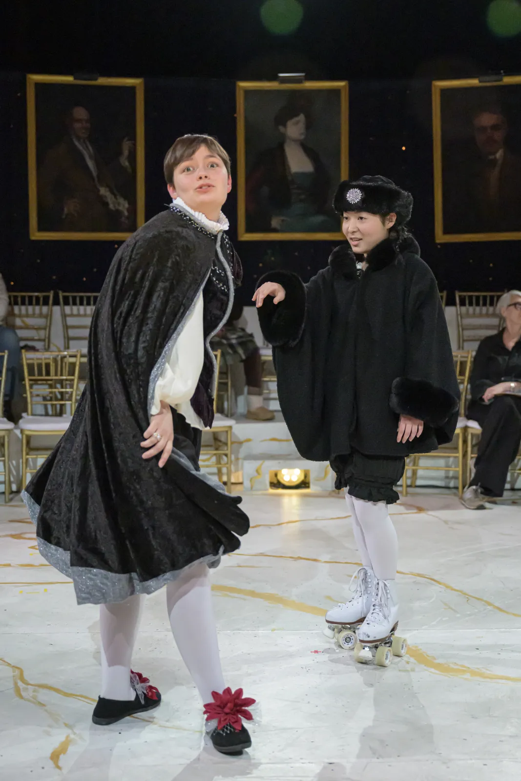 Lizzie Culp looks at the camera with a quizzical expression, and flipping a cape behind her. Kana Nagata looks on from the side, wearing roller skates.