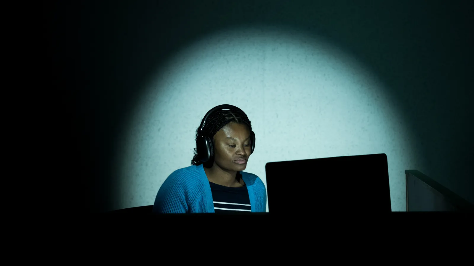 A young person with headphones on looks at their computer. They are in spotlight