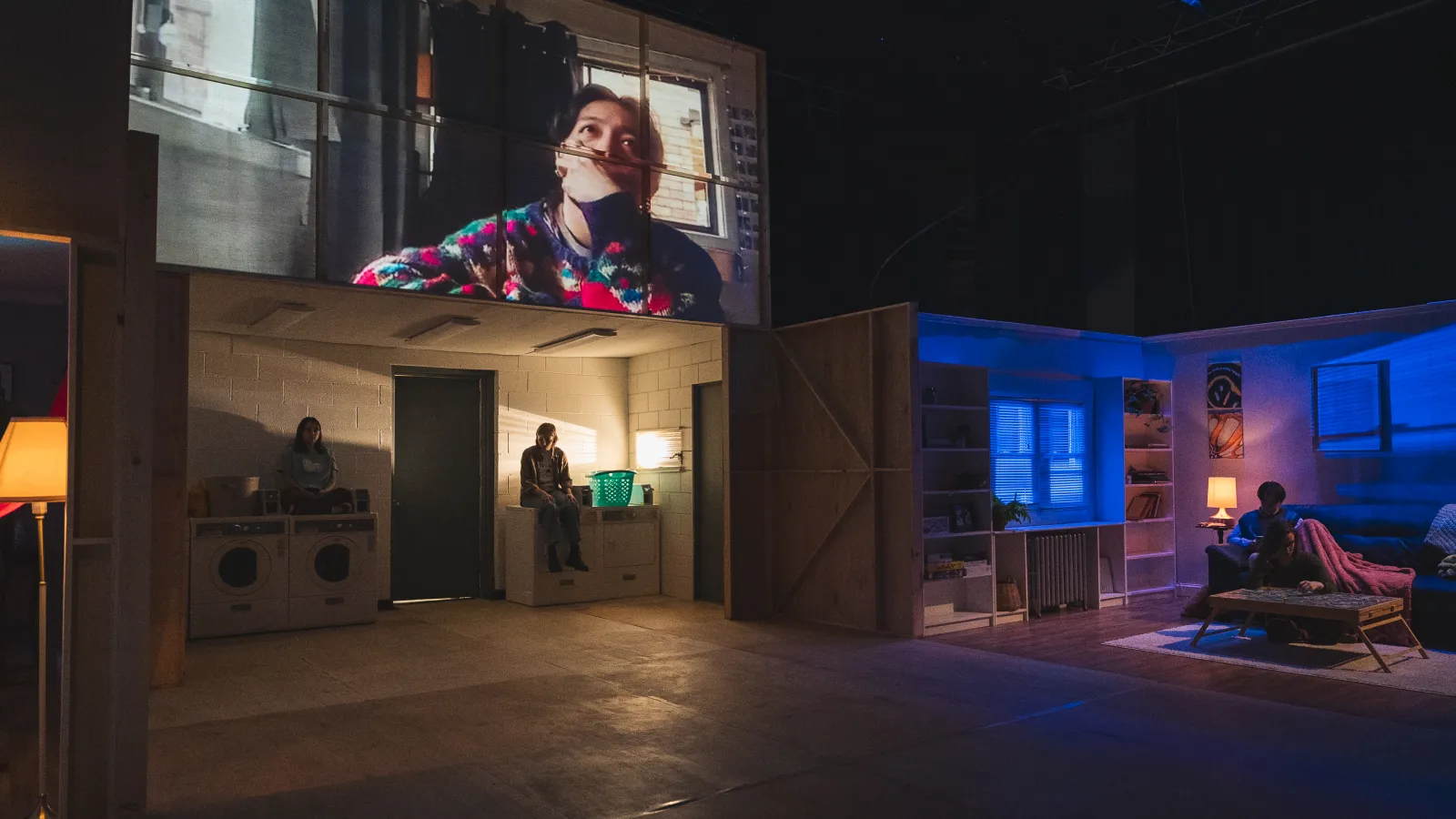 An image from this light says i love you, an Honors Thesis in Directing. A laundry room is wash in warm light, while two individuals sit atop the machines. Above the laundry room is a still from a web-conferencing call featuring an individual in a green and red sweater. They have their hand over their mouth. Across from the laundry room is a living room illuminated in cool blue light. A solitary figure sits on the couch under a blanket. 