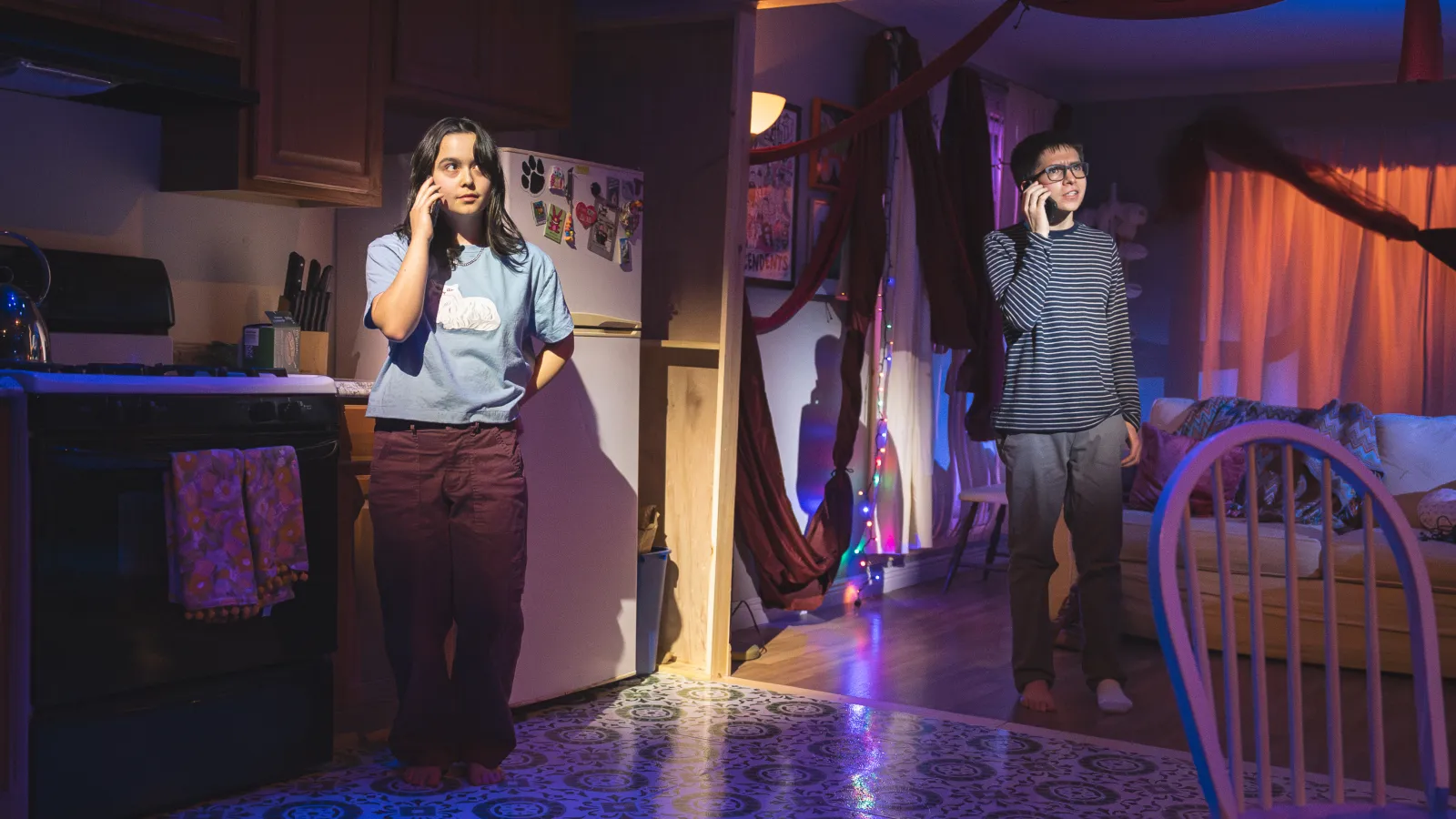 Image from this light says i love you. Two figures stand in two different rooms but are only a few feet apart. They both hold phones to their ears as if in mid conversation. The figure on the left is a femme person in a light blue shirt and maroon pants. They stand in front of their kitchen stove. The performer on the right is masculine appearing, and wears a horizontal striped blue and white shirt. Neither of them are wearing shoes. 