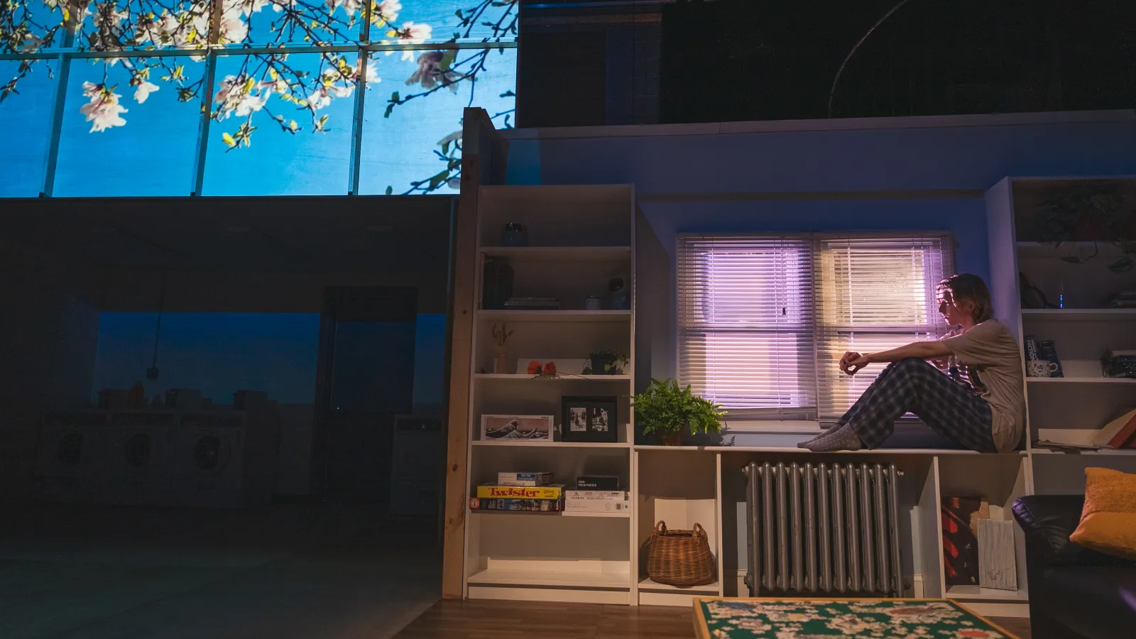 Photo from this light says i love you. A performer sits on top of a white shelving unit in front of horizontal blinds. In the top left of the image, a projection of a tree in bloom can be seen against a blue sky 