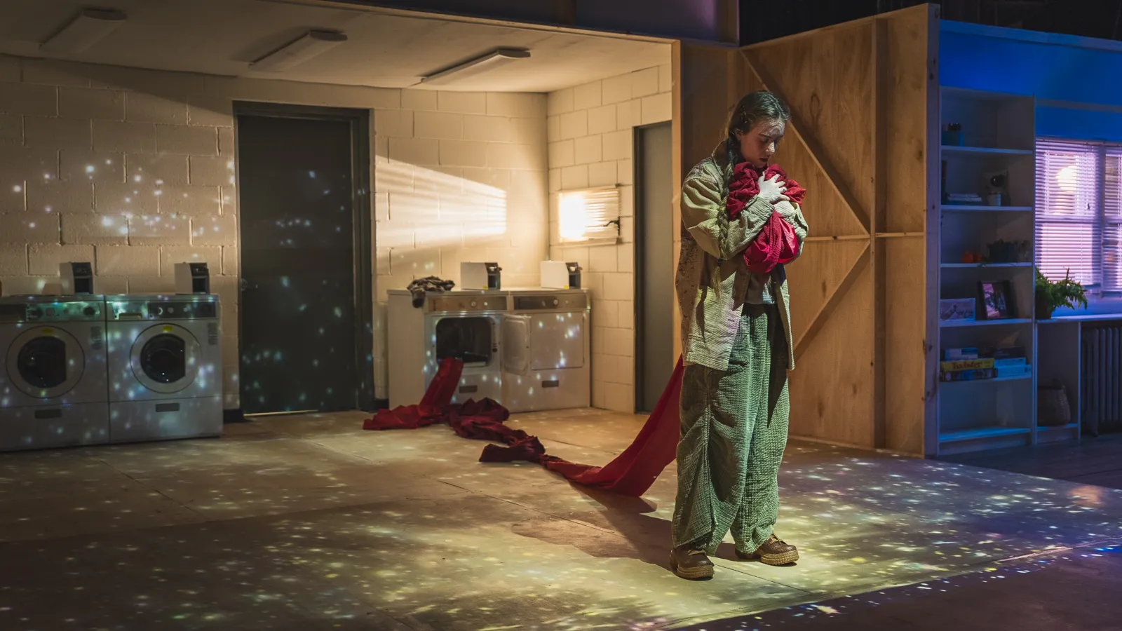 Photo from this light says i love you. A performer stands center stage clutching a red piece of fabric that snakes back into a washing machine upstage. Behind them is a small set of washers and dryers, as if in an apartment or dorm building. The performer is wearing loose fitting jeans and a khaki jacket