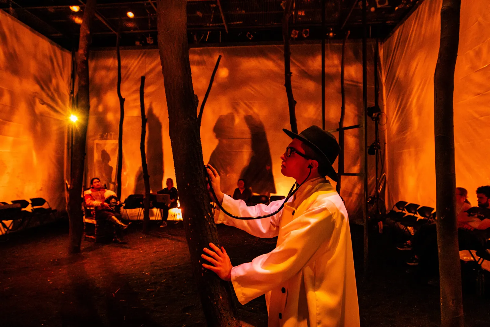 A still from Swarthmore's Woyzeck. A doctor in a dark hat holds a stehoscope up against a tree