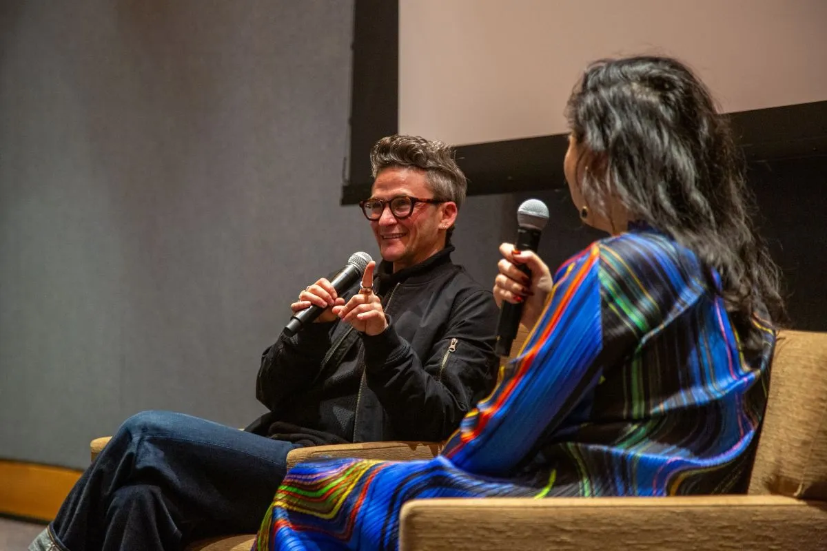 Sam Feder and Dahlia Li sit in mustard colored chairs at the front of the LPAC Cinema in front of the screen. They both hold microphones. Sam is wearing a black shirt, black jacket, and blue jeans. Dahlia is wearing a dress with blue, yellow, red, green, and black wavy vertical stripes