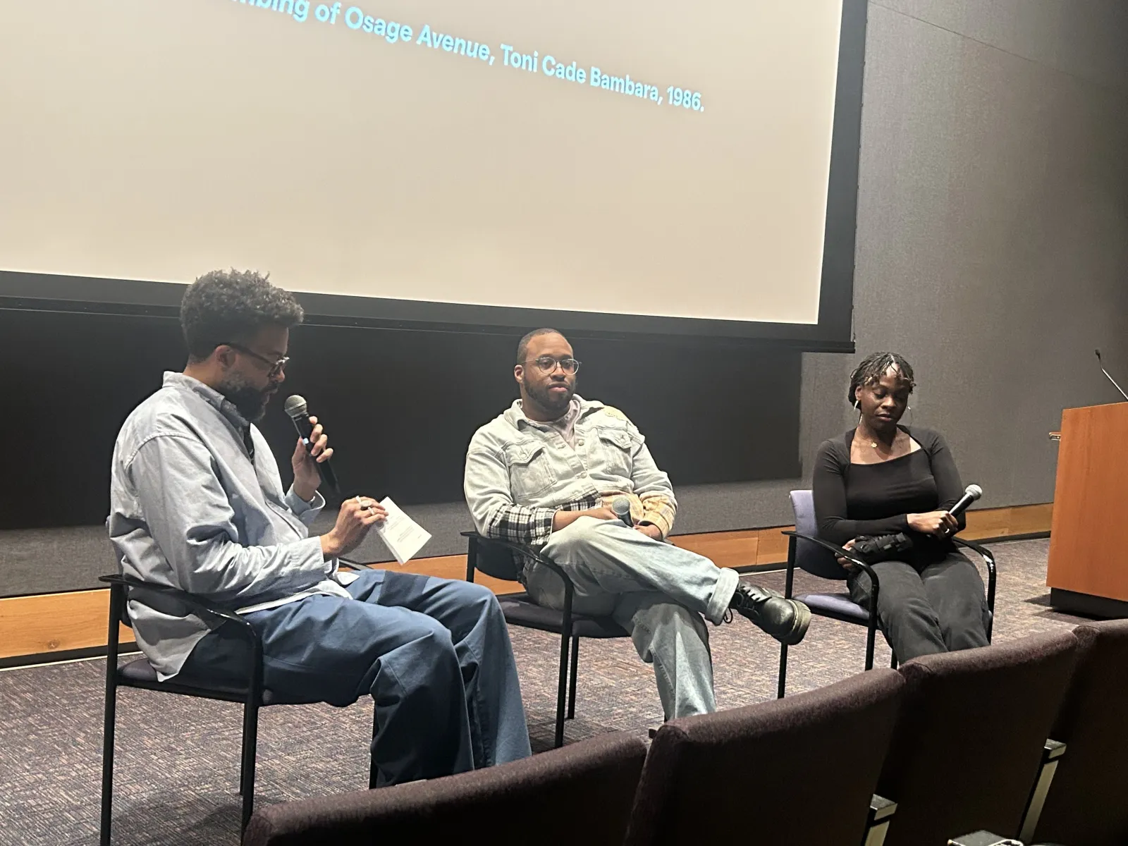 Jamal Batts, Marcellus, and Chisom Chieke sit on chairs at the front of the LPAC Cinema holding microphones. Jamal Batts is holding a piece of paper in one hand and speaking into a microphone held in his other hand