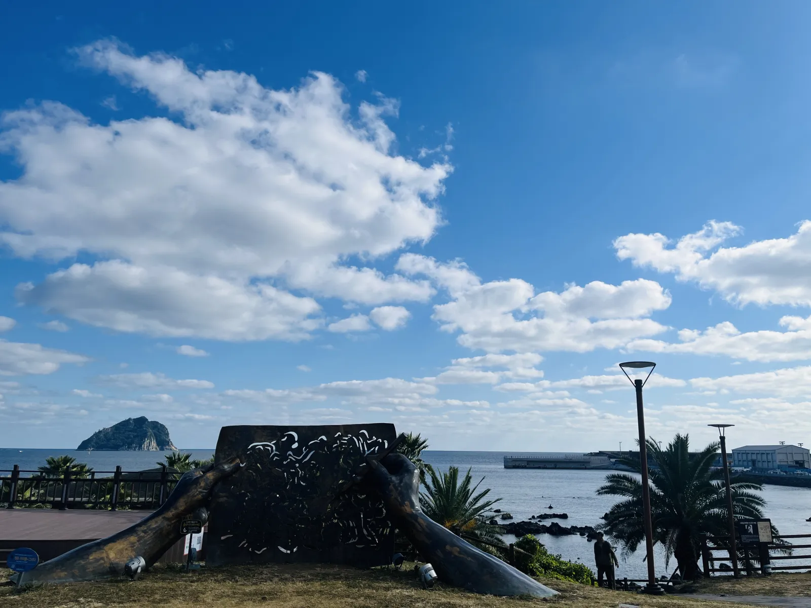 Photo of a view of the ocean and land from Jaguri Park on Jeju Island. A metal sculpture featuring two hands holding and drawing on a sheet of paper, as well as a palm tree are in the foreground. The sky is blue with a few white clouds