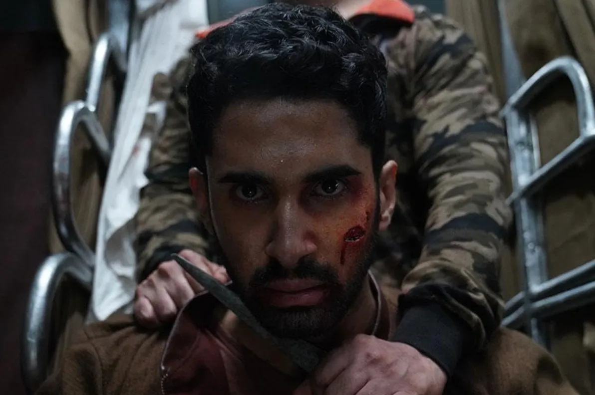 Still from KILL showing a man looking straight forward while another man standing behind him and wearing camo holds a knife against his neck