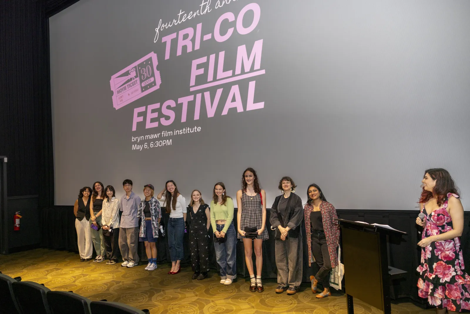 Student filmmakers attend the Tri-Co Film Festival. They stand in front of the screen in a theater at the Bryn Mawr Film Institute. The screen reads Tri-Co Film Festival in pink letters behind them.