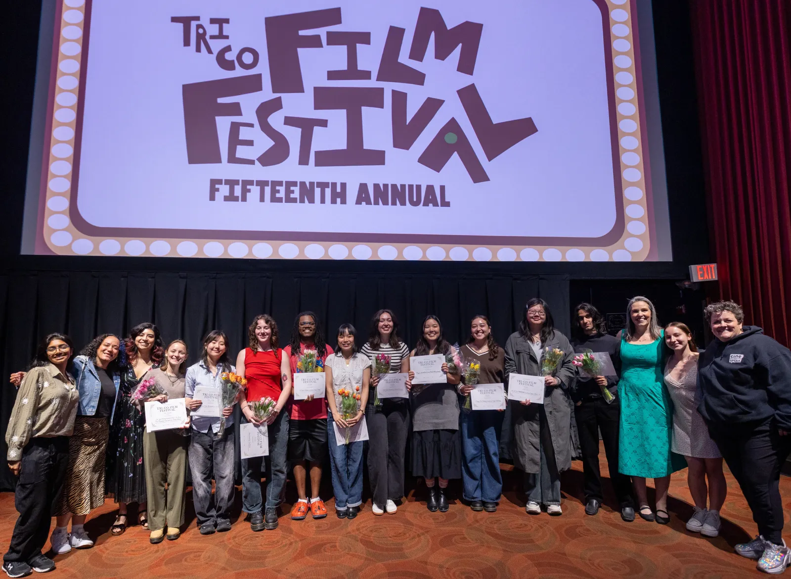 Student filmmakers, Tri-Co Film Festival interns, Tri-Co Film Festival judges, and Ellen Sanders and Kristal Sotomayor smile for a photo at the front of a screening room at the Bryn Mawr Film Institute. The students each hold a bouquet of flowers and Tri-Co Film Festival Fifteenth Annual is projected onto the screen