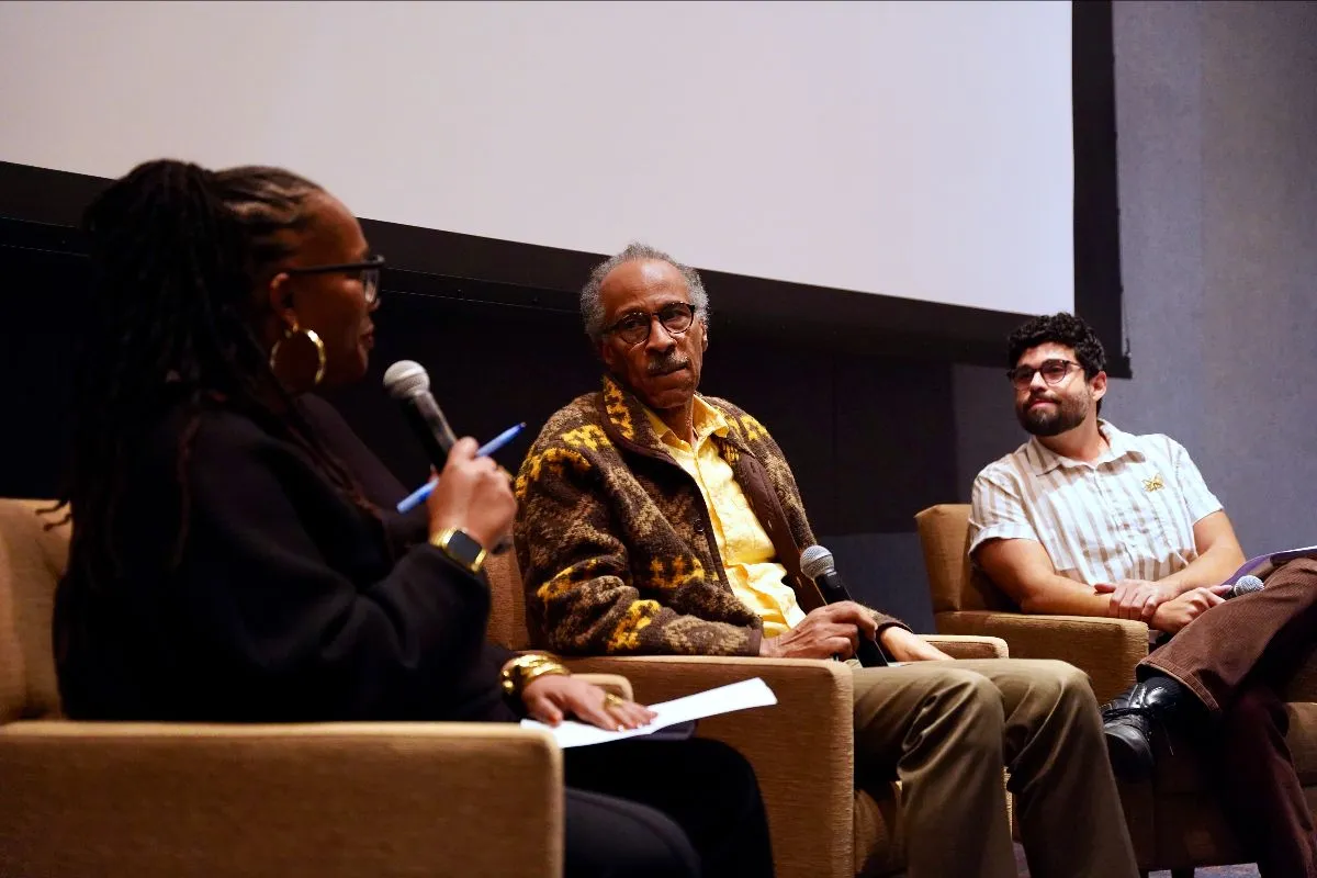 Nina Johnson, Louis Massiah, and Conor “Coco” Tomás Reed hold mics and sit in yellow chairs in front of the screen in the LPAC Cinema. Nina Johson is wearing black pants and a black shirt, Louis Massiah is wearing brown pants, a yellow shirt, and a yellow and brown jacket, and Conor “Coco” Tomás Reed is wearing brown pants and a white and tan striped shirt with a gold butterfly pin