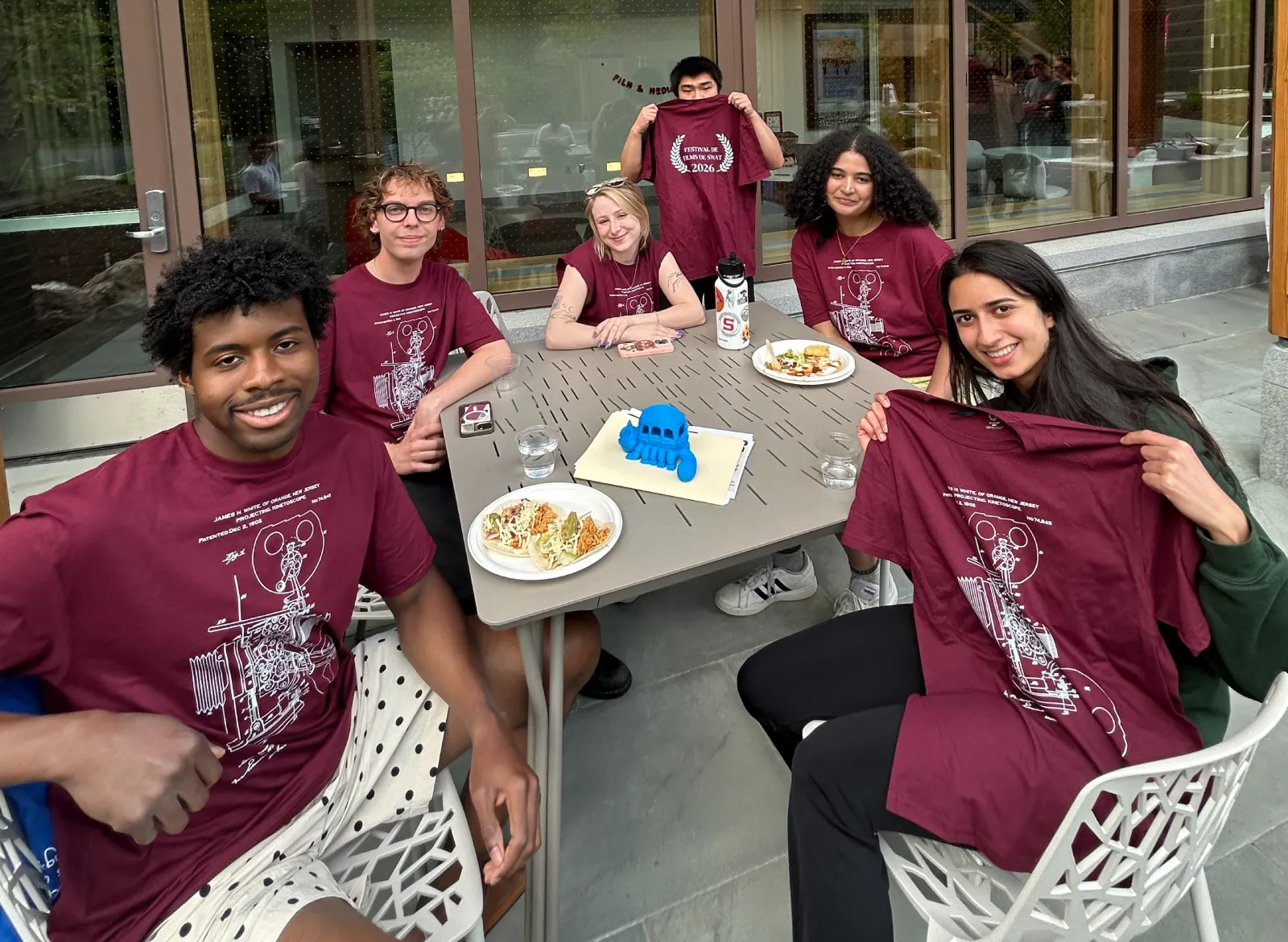 Six Film & Media Studies seniors at the FMST spring picnic smiling and holding up their class shirts. The shirts are garnet and read Festival De Films De Swat.