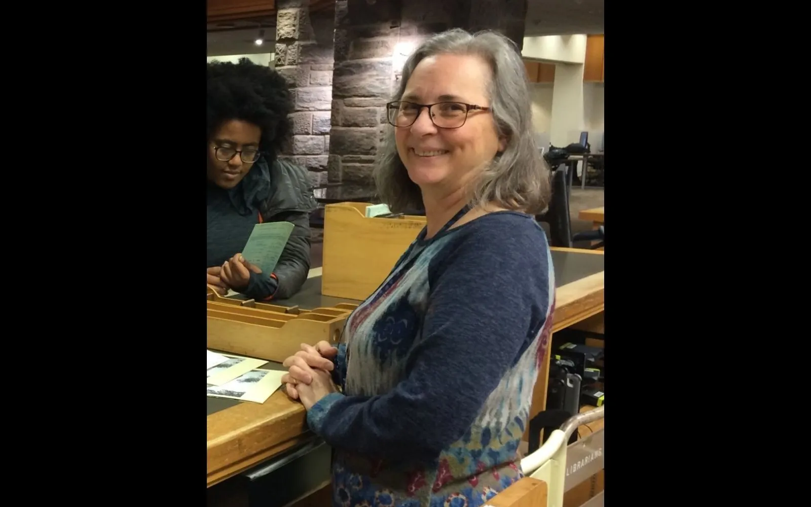 A woman with shoulder-length grey hair and glasses wearing a dark top smiles over her shoulder at the camera.