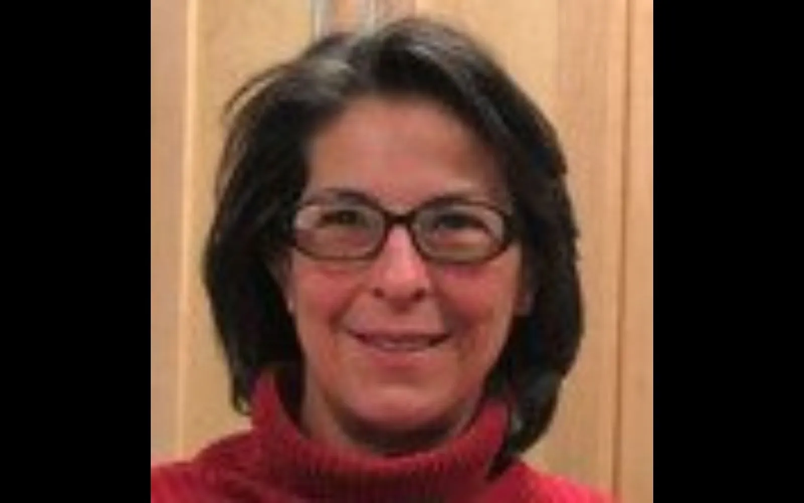 Close shot of a woman with shoulder length grey hair wearing glasses and a red turtleneck, smiling at the camera.