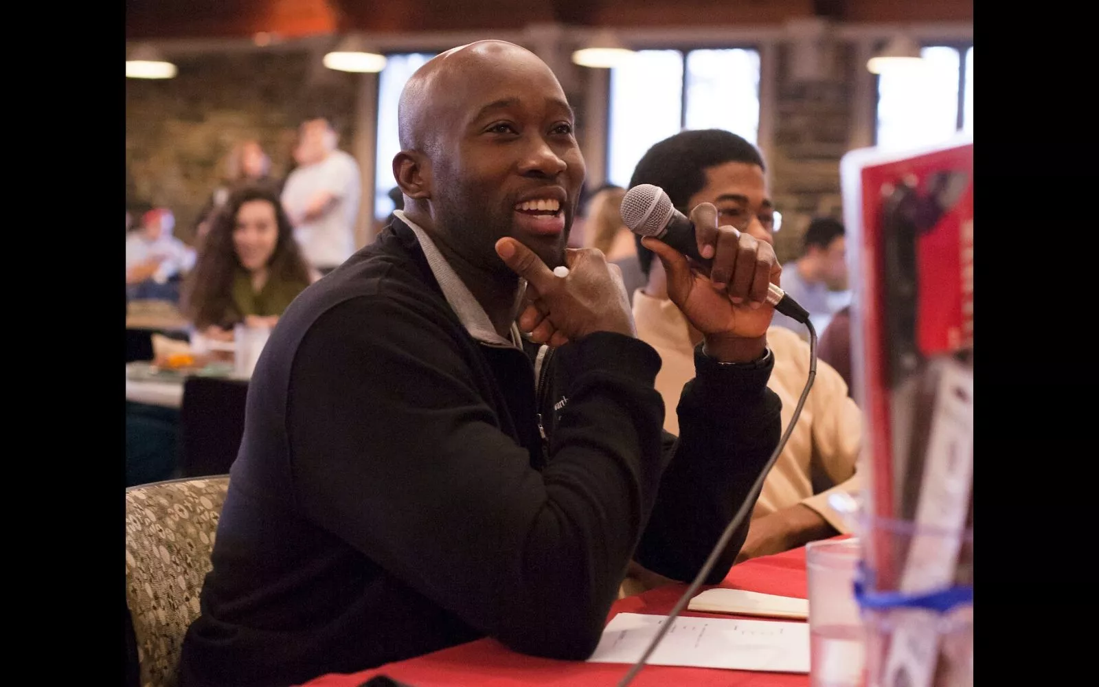 Smiling man with a shaved head wearing a black top sits at a table with a microphone