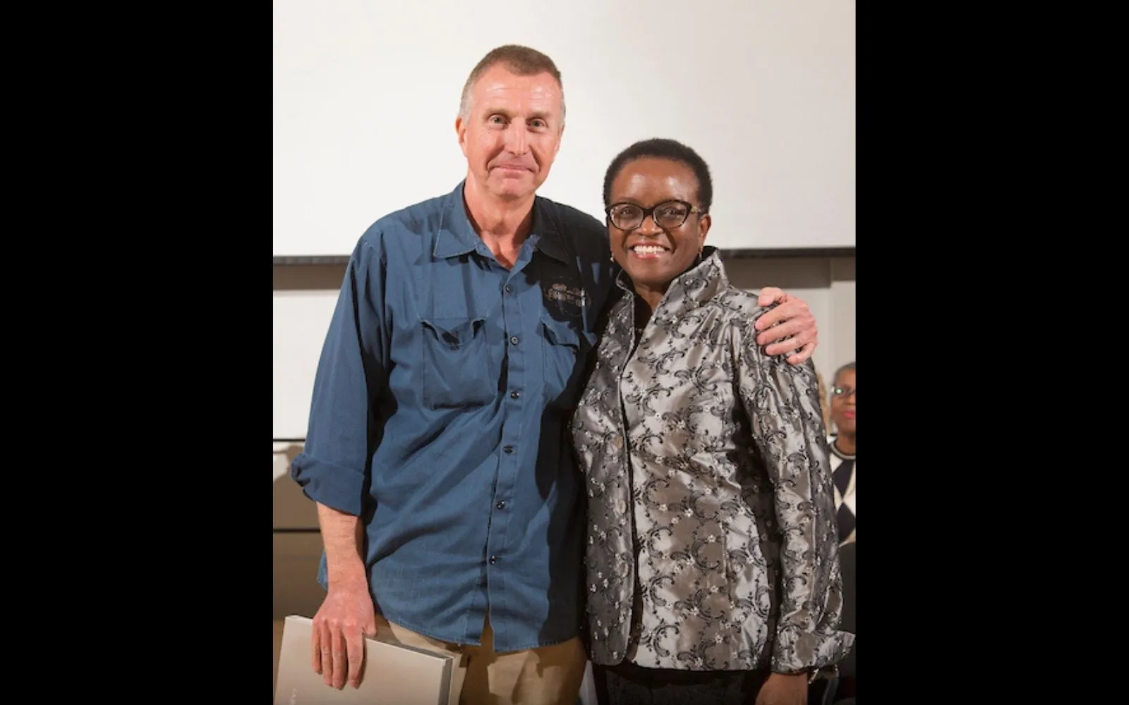 A tall man with close cropped grey hair wearing a blue cargo shirt stands next to a woman with close cropped dark hair and glasses, wearing a patterned grey jacket. Both smile at the camera.
