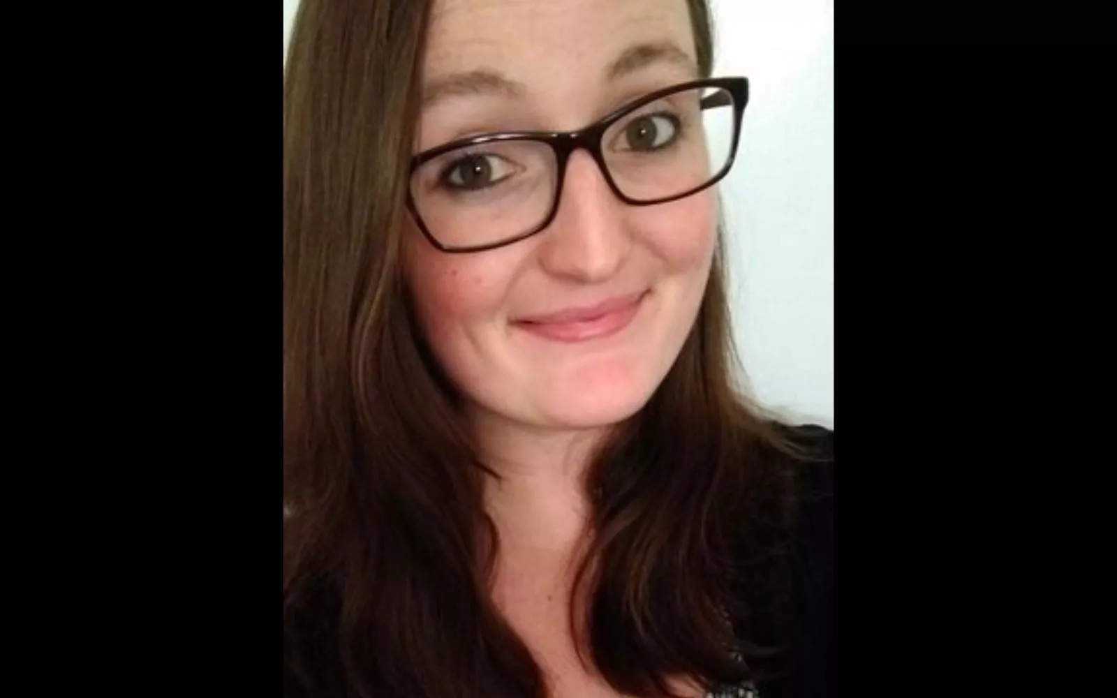 Headshot of a woman with long brown hair and black glasses grinning at the camera