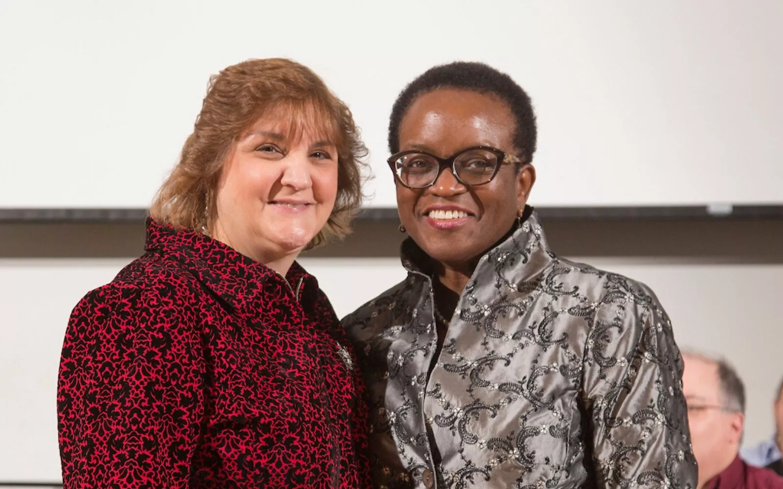  Woman with short curly brown hair and a dark red top stands next to College President Val Smith, who has short hair and glasses.