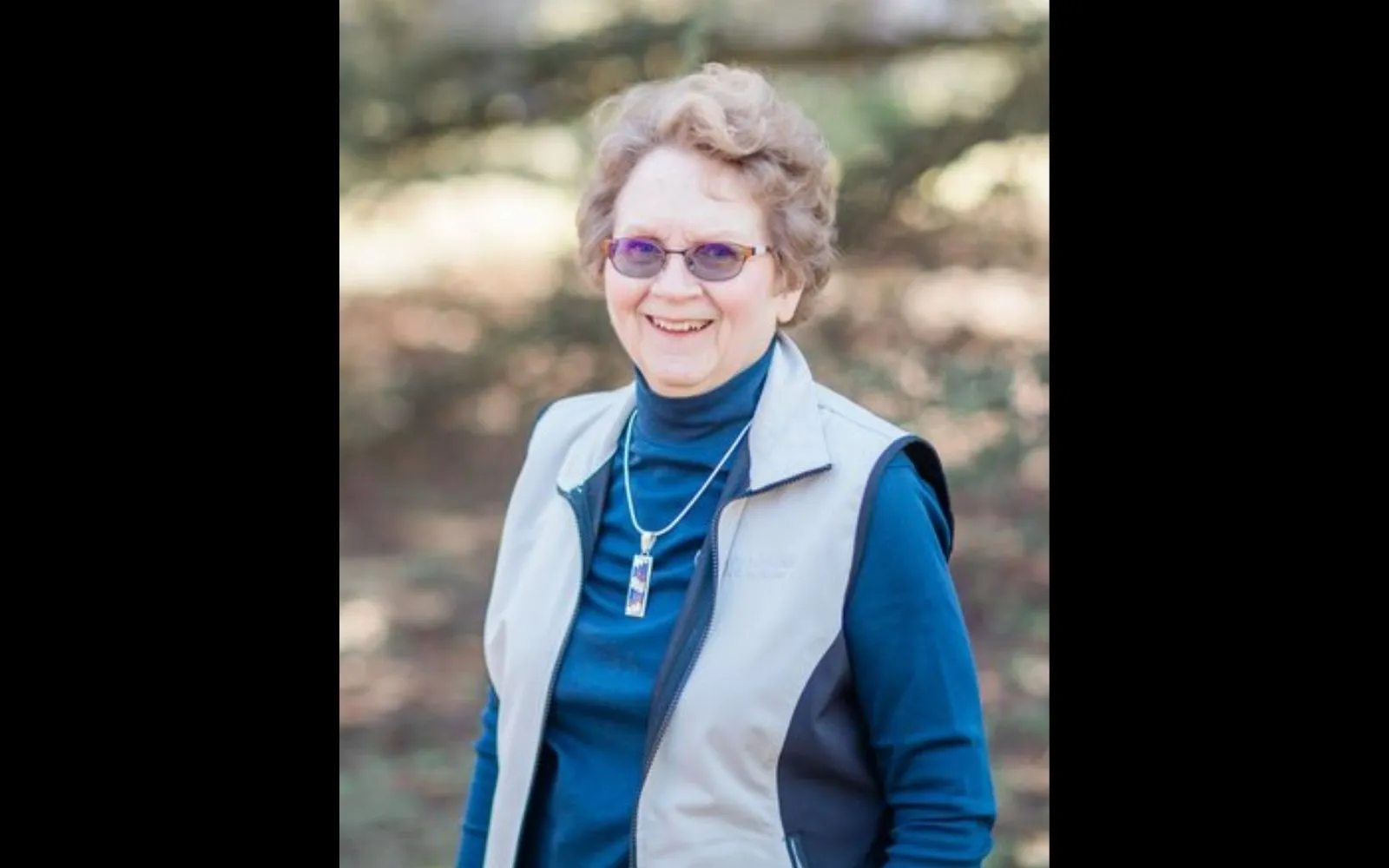 A woman with short grey hair and glasses stands outside in a blue top and white vest, smiling at the camera.