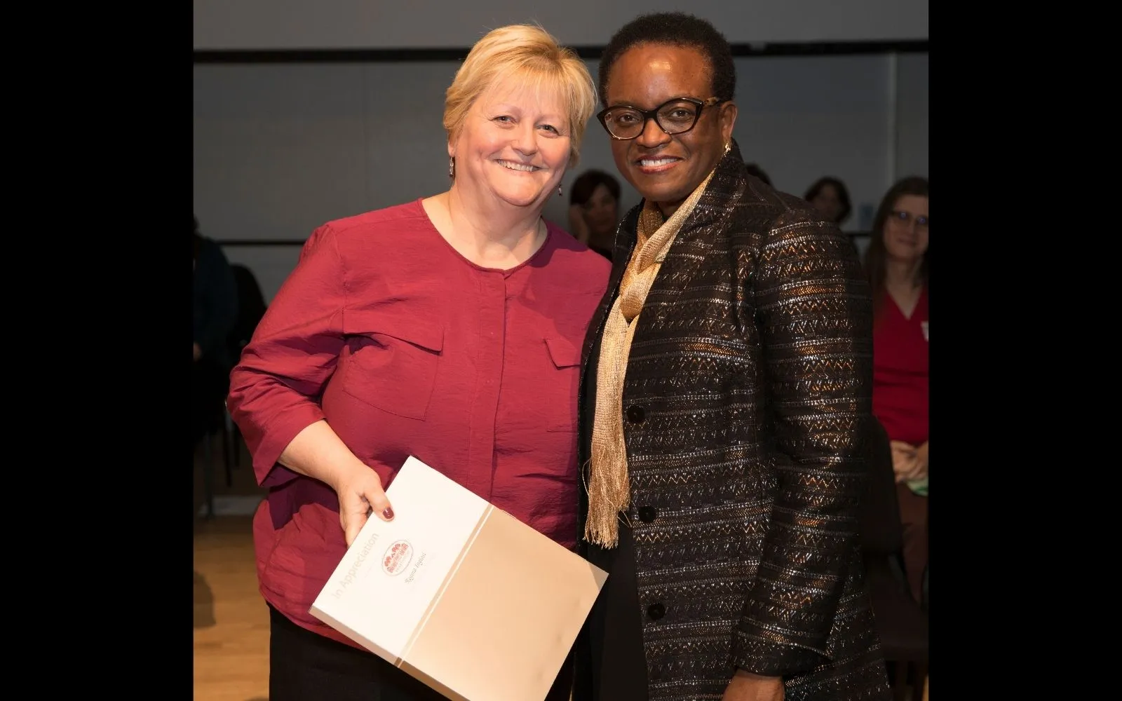 Two woman stand beside each other and embrace, smiling at the camera. The woman on the left wears red top and has short blonde hair. The woman on the right has close cropped hair and wears glasses and a long dark jacket.