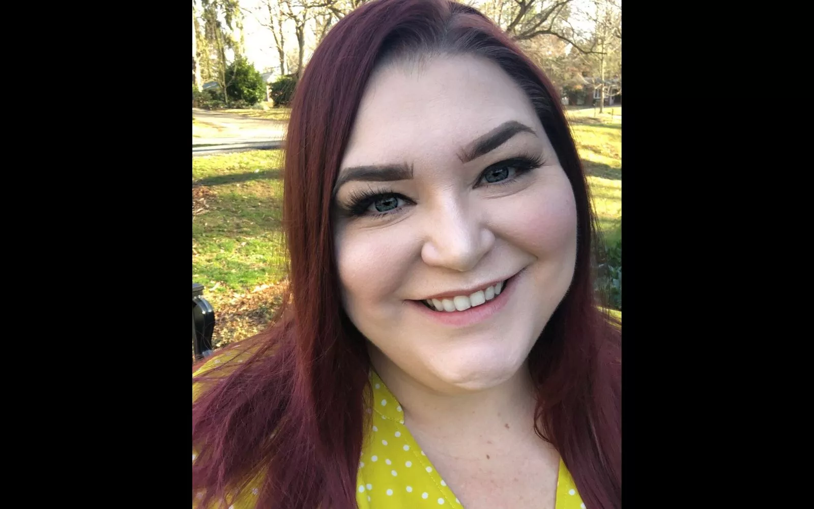Woman with long burgundy hair and a bright yellow top smiles at the camera