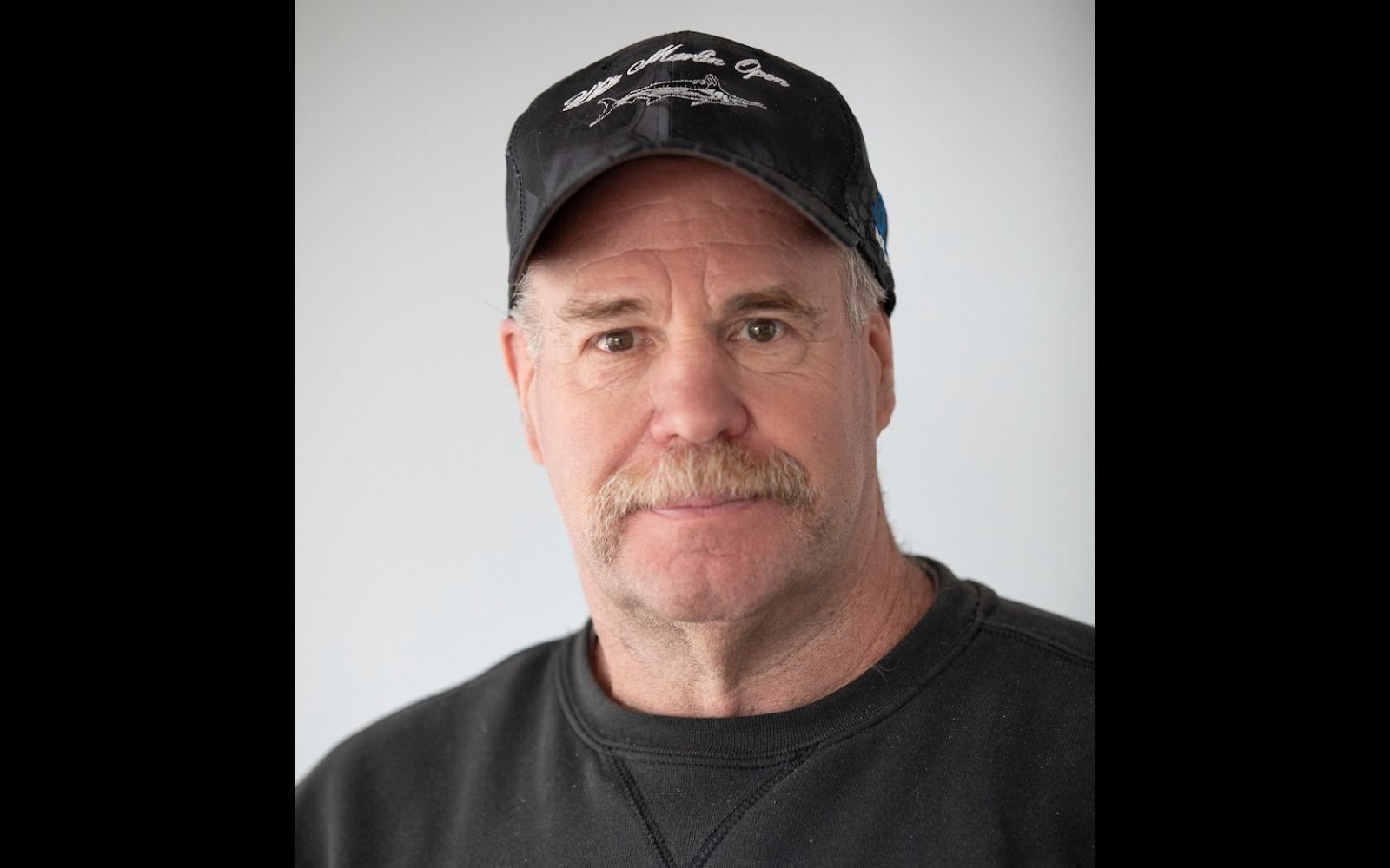 Head shot of a man with a white mustache wearing a black baseball cap and black tee shirt.