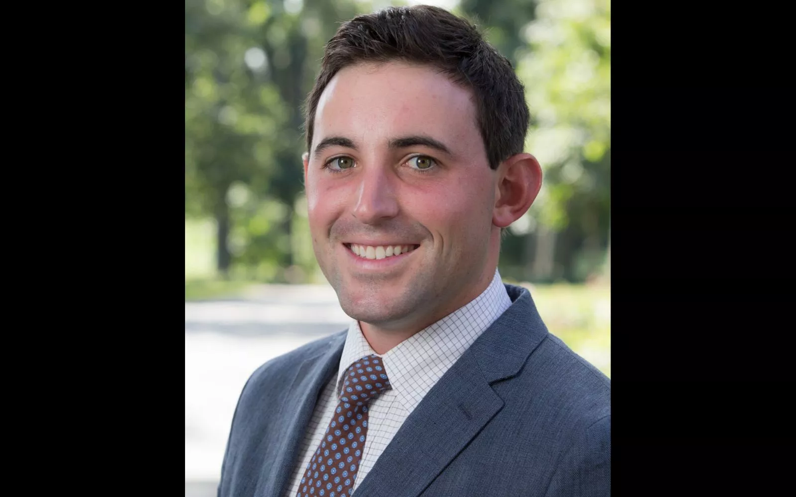 Headshot of man with short brown hair wearing a grey suit, white shirt, and dark tie.