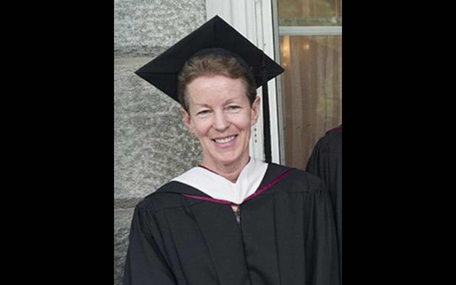 A woman with close cropped hair, wearing graduation regalia, smiles at the camera