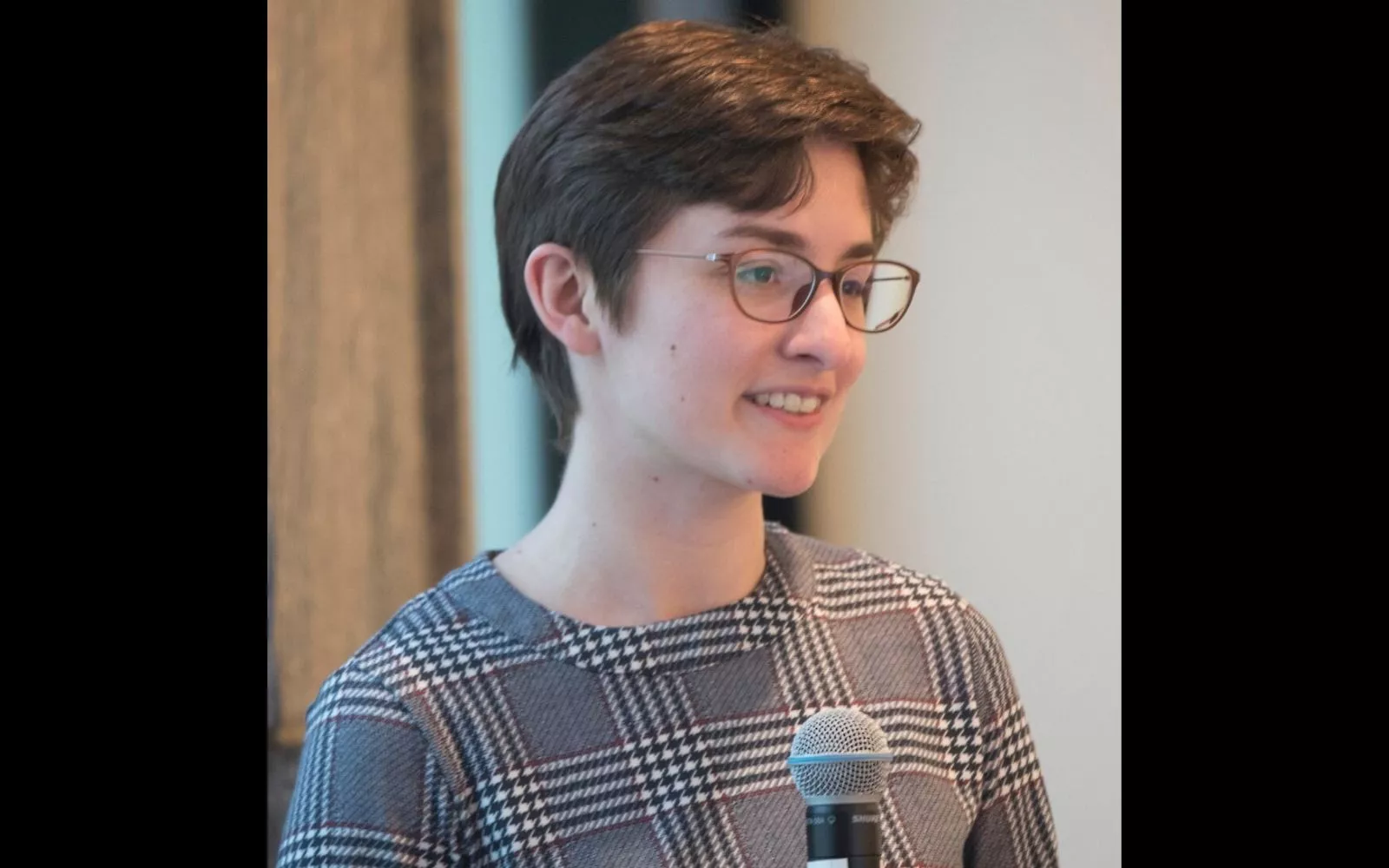 Woman with short brown hair and glasses wearing a checkered dress holding a microphone.
