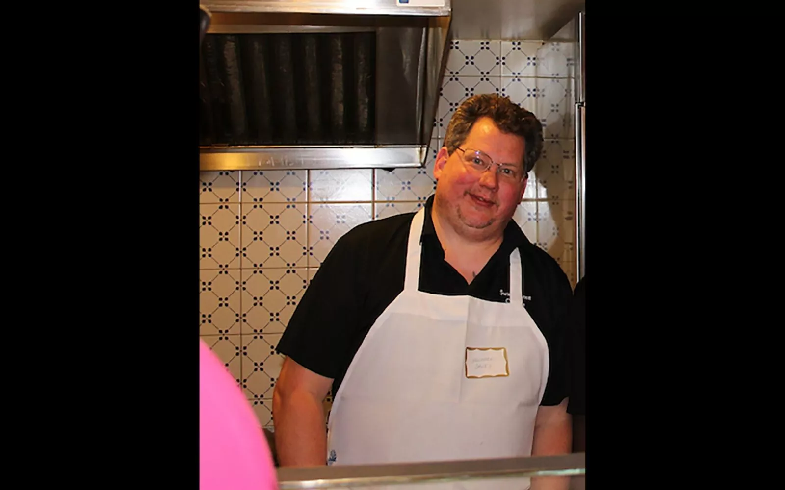 Man with glasses and brown hair wearing a white apron and standing in an industrial kitchen.